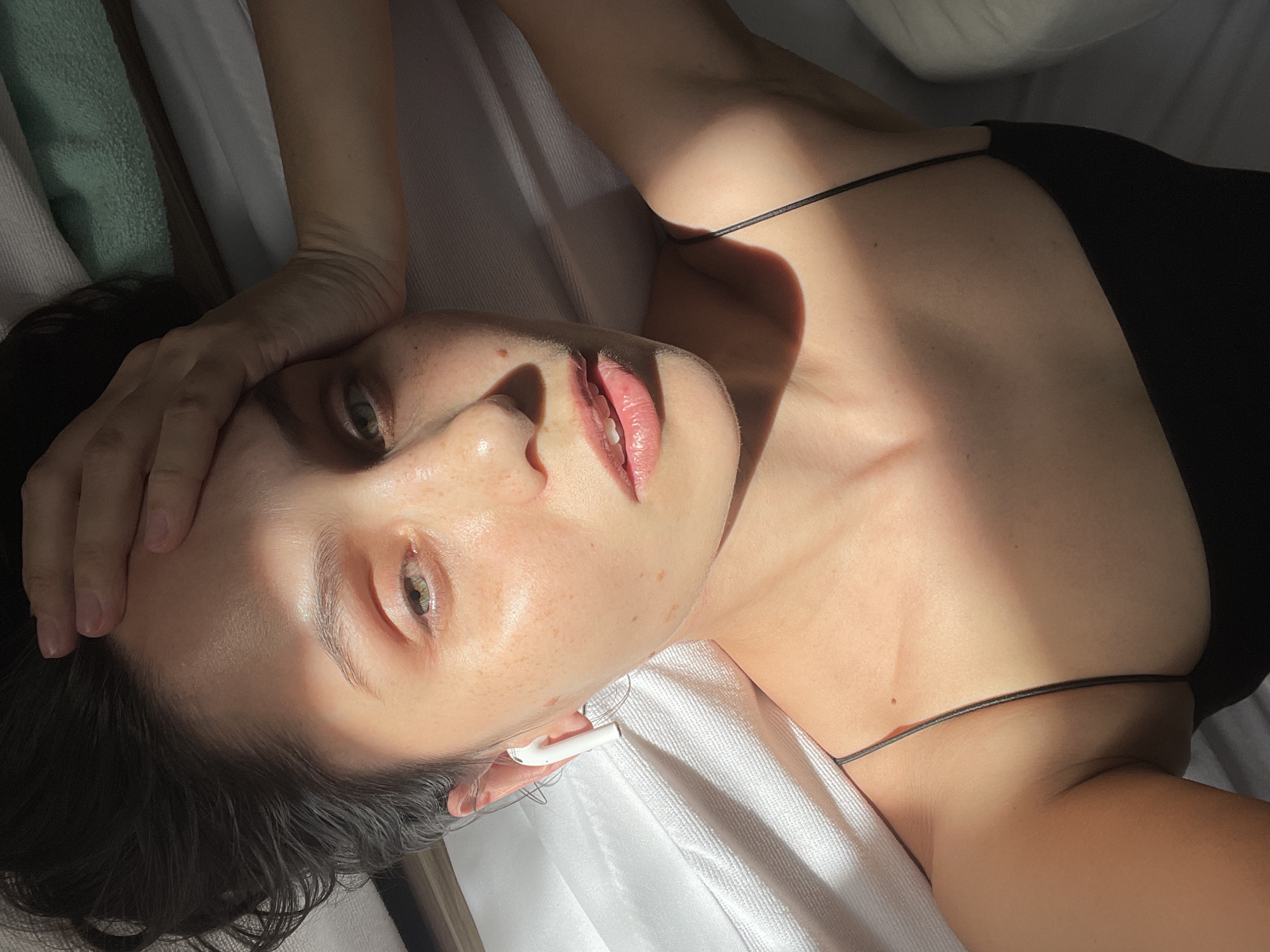 A woman lies on a white surface. She is wearing a black spaghetti strap top and has an earbud in her right ear. She is fair-skinned with dark hair. Her left hand is near her forehead. Light is shining across her face and chest. The location is not discernible from the image.