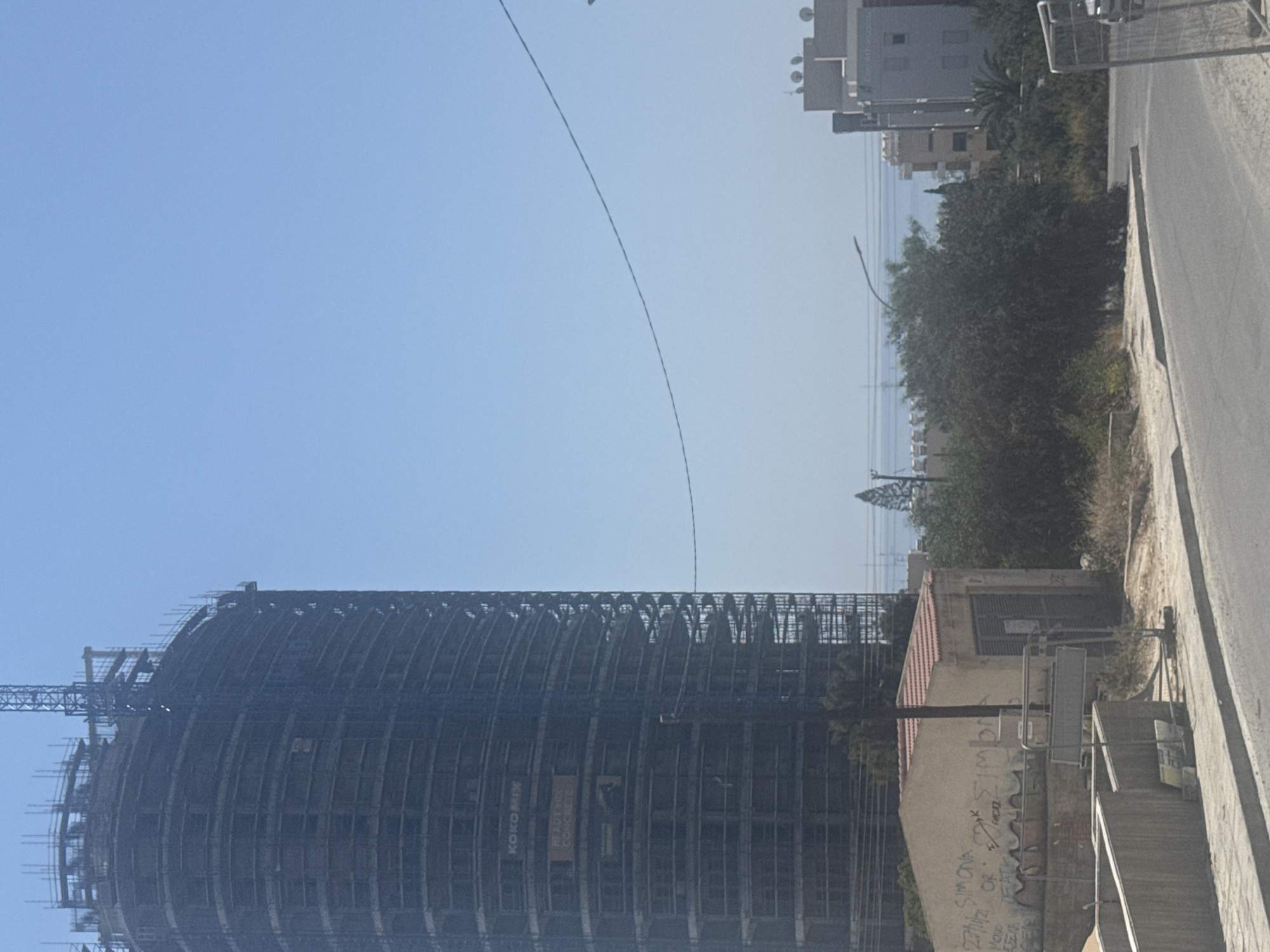 A tall, circular building under construction, covered in scaffolding, dominates the left side of the image. The building has a crane at its summit. A sign reading "KOKOMIX READY MIX CONCRETE" is visible on its facade. In the foreground, a small, light-colored building with a red tile roof is covered in graffiti, including what appears to be "SIMONA" and "OR". A street runs in front of this building, and to the right, other buildings, including a taller, modern structure, are visible in the background. The scene is set against a clear blue sky, with hints of the sea visible in the distance between buildings. The environment suggests an urban or developing area in Agios Tychona Community, Cyprus.
