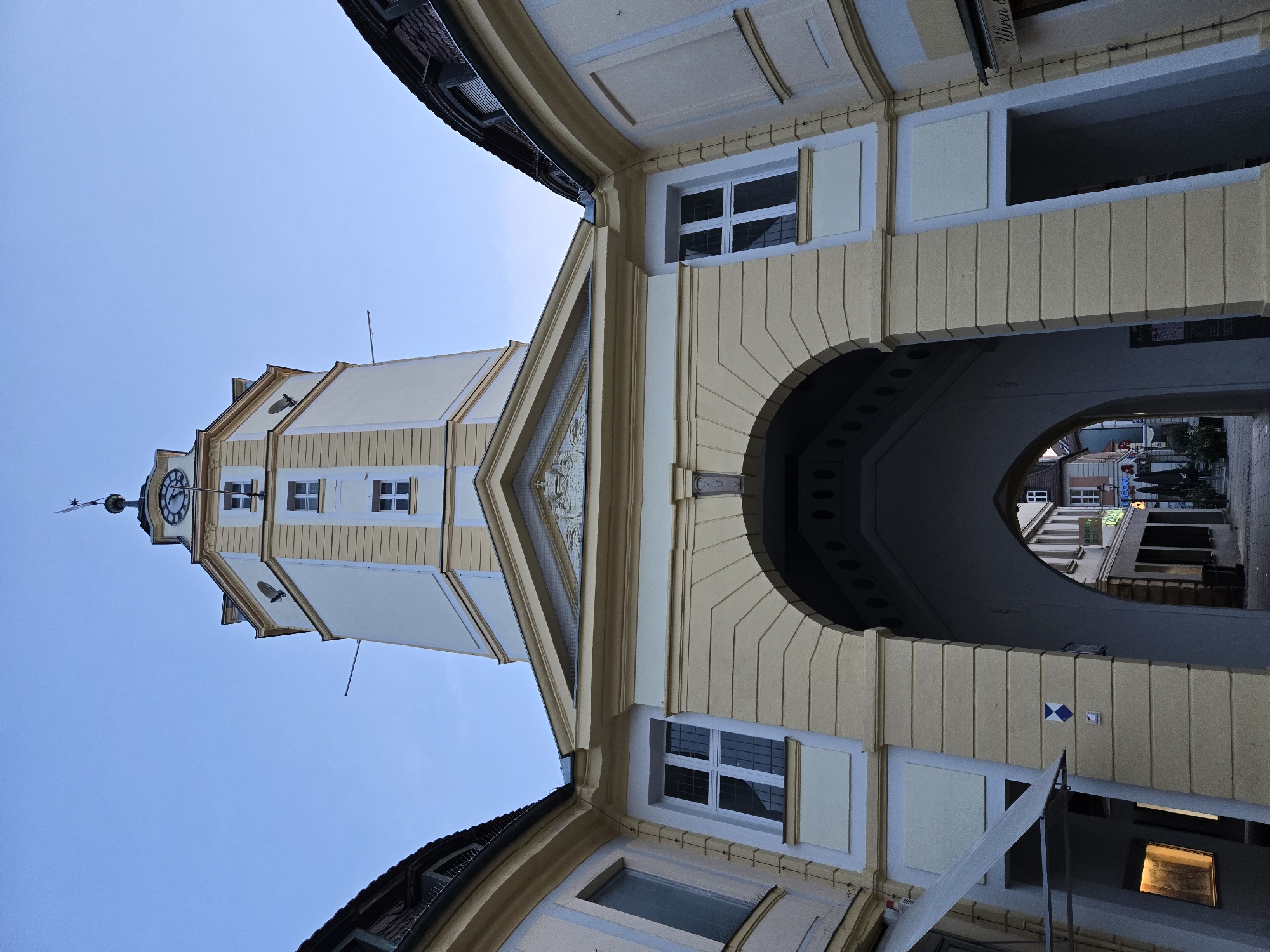 The image shows a low-angle view of the Ansbach city hall tower and its adjoining archway in Ansbach, Germany. The tower is tall and octagonal, painted in cream and yellow with white accents. It features a clock face on one side and several rectangular windows. At the very top, a weather vane is visible. The archway, which leads into a street, is large and curved, supported by substantial pillars. The buildings flanking the archway have curved, tiled roofs and are painted in pale yellow and cream. Several windows with white frames are visible on these buildings. The sky above is a clear, pale blue. The street visible through the archway shows more buildings with shops, awnings, and some outdoor seating. The overall scene suggests an urban setting, likely in a historic European town.