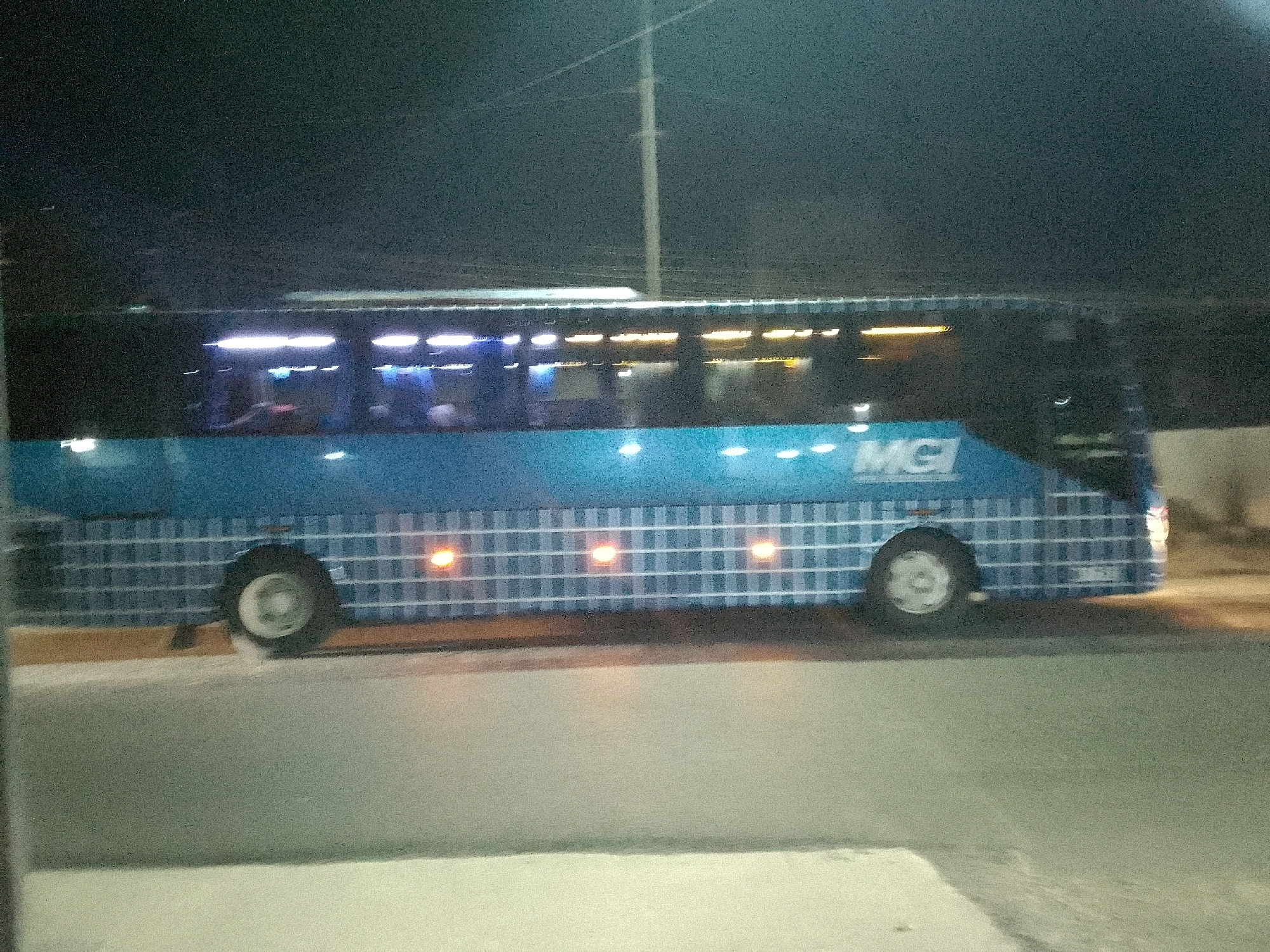A blue bus with a white checkered pattern is parked at night on a paved road. The bus has tinted windows with interior lights visible. The word "MGI" is visible on the side of the bus. The scene appears to be in an urban or semi-urban setting, possibly a bus terminal or a roadside stop. The lighting conditions suggest it is dark outside, with artificial lights from the bus and the surroundings illuminating the scene. The image is slightly blurry, indicating potential motion blur or low light photography. There are no people clearly visible in the image. The bus is the primary subject, and its branding suggests it is part of a transportation service. The overall environment is a dimly lit road with the bus as the focal point. The location is identified as Gunungmasigit, Indonesia.