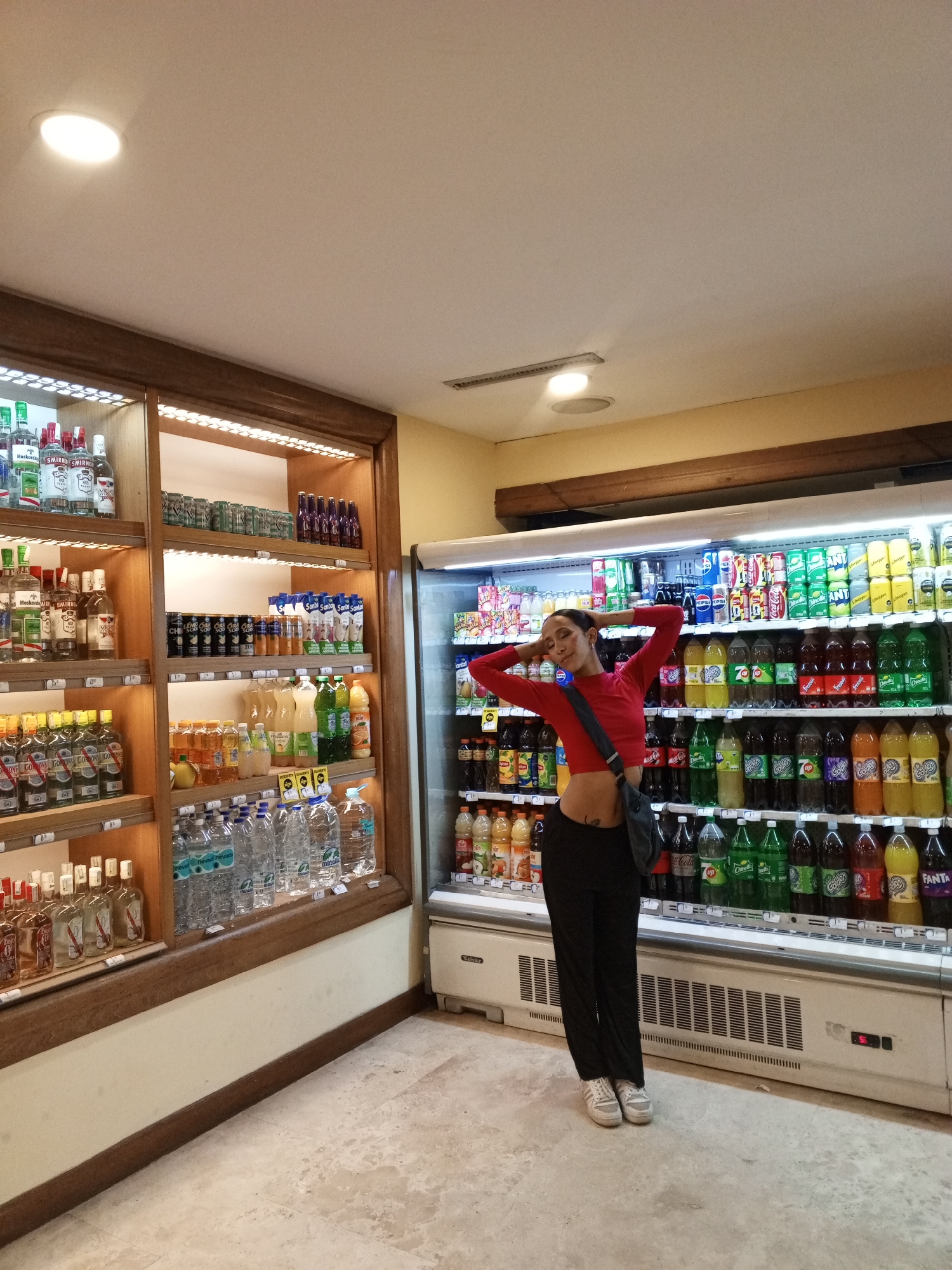 A young woman poses in a convenience store aisle in Porlamar, Venezuela. She wears a red, long-sleeved crop top, black pants, and sneakers, and has a gray shoulder bag. Her hands are behind her head. To her left is a wooden shelving unit stocked with liquor and beverages. To her right is a refrigerated display case filled with soda and juice. The floor is light-colored tile. Overhead are recessed lighting fixtures.