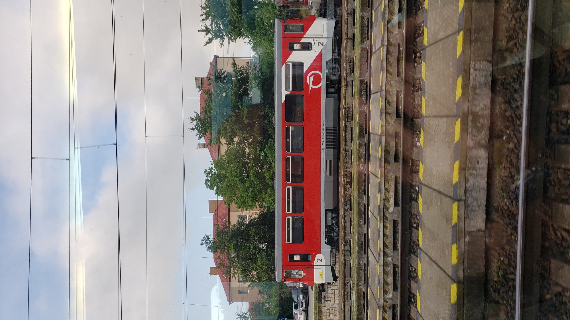 A red and white train car is stopped at a station in Margecany, Slovakia. The train car has a number "2" displayed on both ends. The station platform is visible with yellow and black hazard stripes. In the background, there are buildings with red roofs and green trees. The sky is partly cloudy.