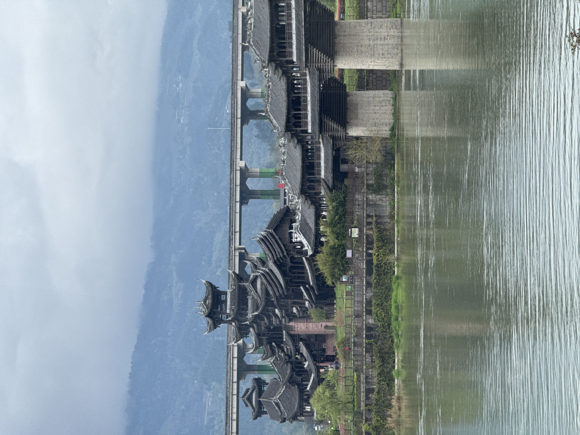 The image shows a horizontal view of a landscape. The right side displays a body of water with visible ripples. Adjacent to the water is a green space with trees and grass. A structure with a dark roof sits along the green space. To the left, a multi-tiered pagoda-style building extends into the water. A bridge runs parallel to the pagoda, with concrete supports visible. In the far left background, a mountain range appears under a cloudy sky. This is a scene in Zhuoshui, China.
