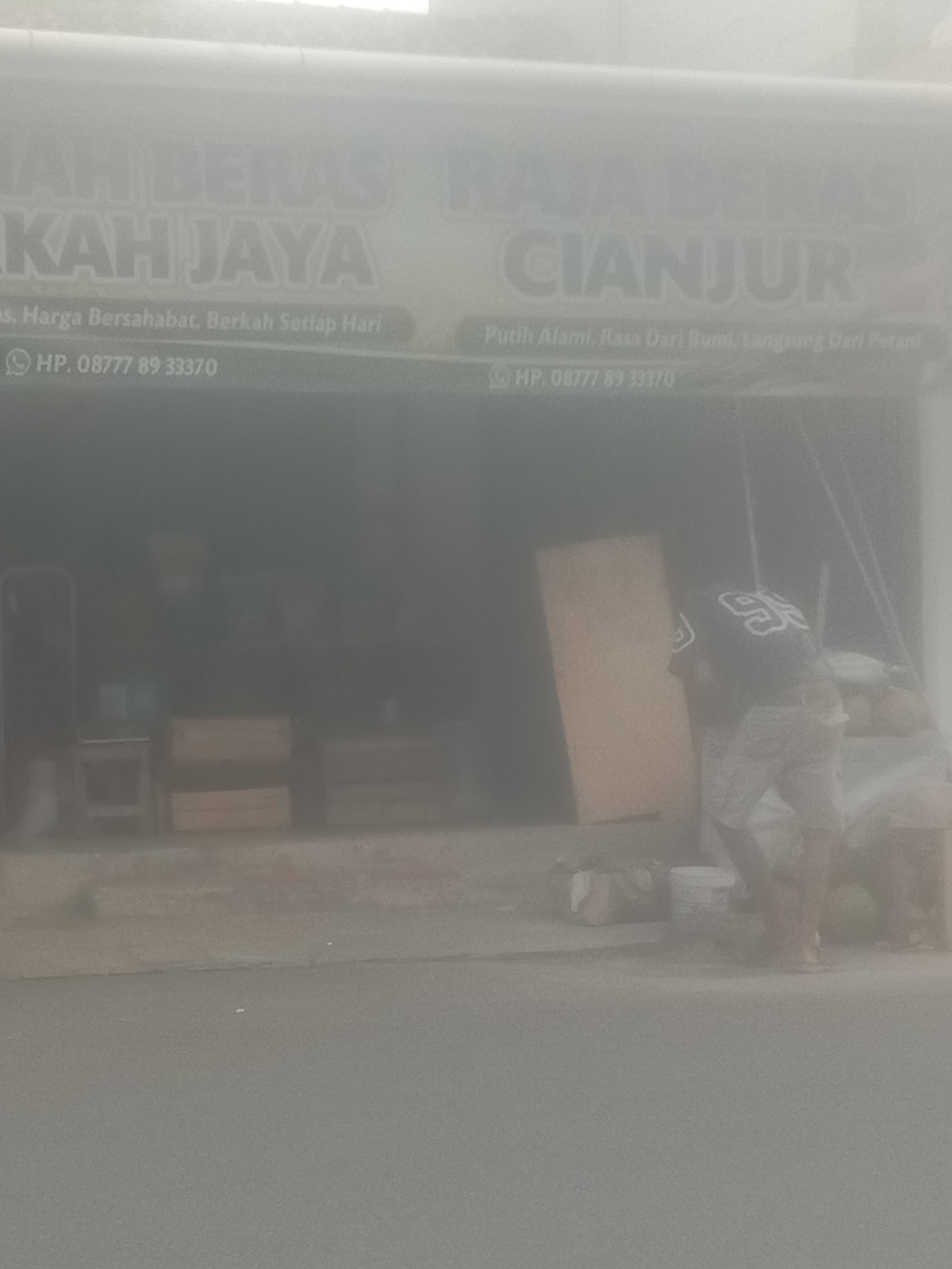 The image depicts a street-level view of a shop in Bojong, Indonesia, with a focus on its facade and a small portion of the street. The shop's signage, partially obscured by glare and atmospheric haze, displays "RAJA BERAS KAH JAYA" and "CIANJUR" in large, bold letters. Below these main titles, smaller text reads "HP. 08777 89 33370" and "HP. 08777 89 33370," suggesting two different points of contact or possibly a repeated contact number. Another line of text on the left side, partially visible, appears to say "as, Harga Bersahabat, Berkah Setiap Hari," which translates to "as, Friendly Prices, Blessings Every Day." On the right side of the shop's interior, a person is visible wearing a black t-shirt with the number "95" in white on the back and patterned shorts. They appear to be engaged in some activity involving a large, flat, light-colored object, possibly a piece of wood or a large sign, and are facing away from the camera. In the background inside the shop, there are wooden crates stacked, and some equipment like buckets. The street in front of the shop is paved and shows signs of wear. The overall scene is slightly hazy, suggesting it might be a dusty or humid environment.