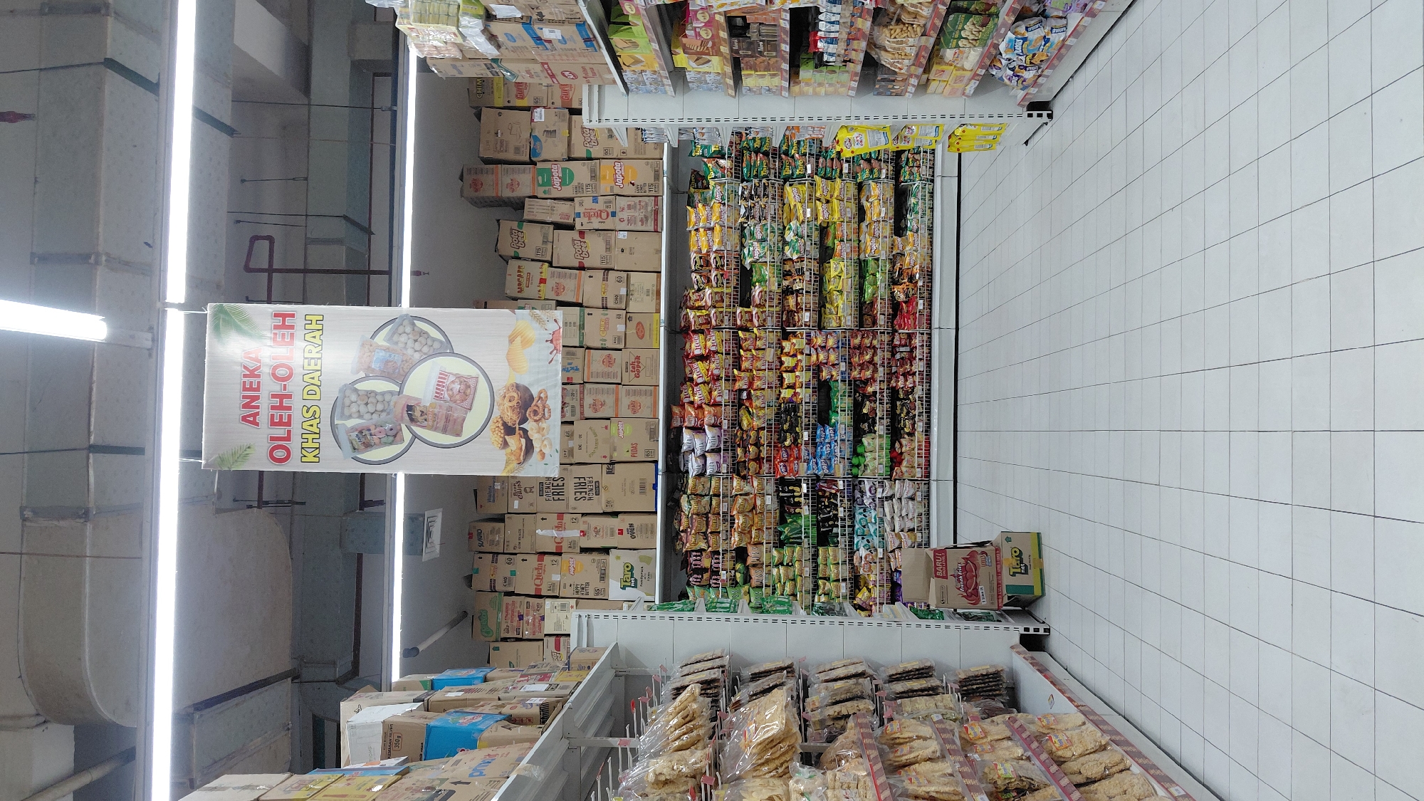 The image shows an aisle in a supermarket in Purwokerto, Indonesia. The aisle is filled with shelves of snacks and other food items. A large banner hangs from the ceiling, advertising "Aneka Oleh-Oleh Khas Daerah," which translates to "Assorted Souvenirs from Various Regions." The shelves are stocked with a wide variety of packaged goods, including chips, crackers, cookies, and candy. The floor is tiled with white squares. The lighting in the supermarket is bright, with fluorescent lights illuminating the products. The overall impression is one of a well-stocked and organized supermarket.