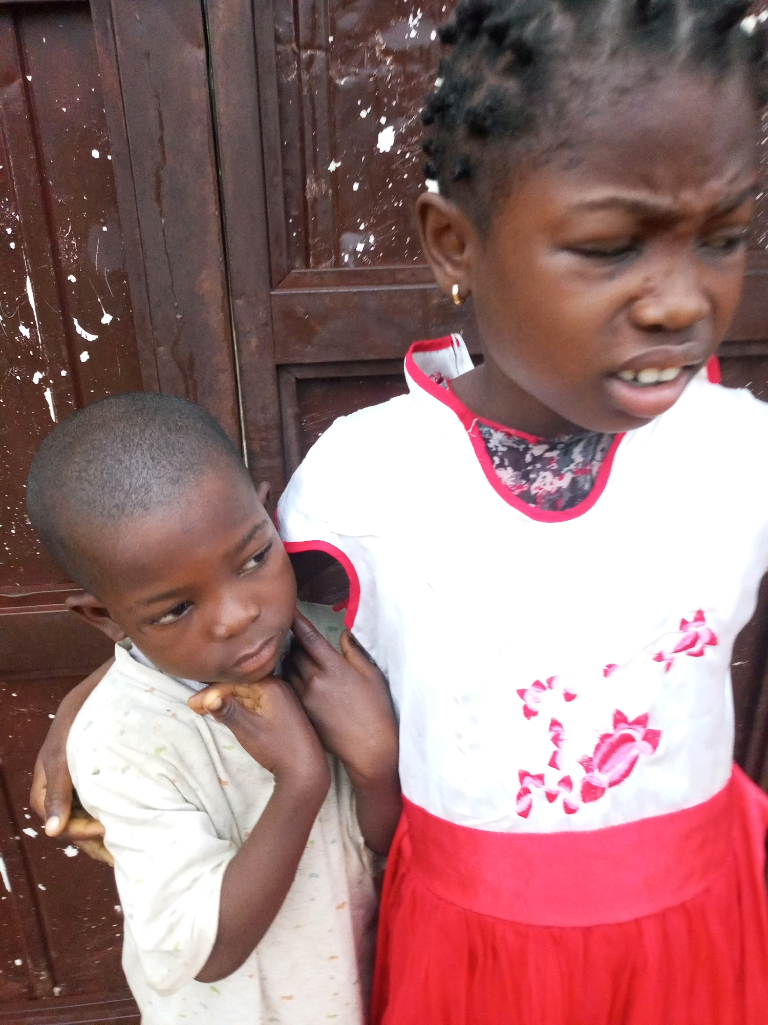 Two young Black children stand in front of a dark brown wooden door speckled with white paint. The child on the left, a boy with a shaved head, wears a light-colored t-shirt with a subtle pattern of small, colorful dots. His head is tilted down, and his hands are clasped in front of him. The child on the right, a girl with braided hair, wears a white top with red trim around the neckline and a red skirt. She has a gold hoop earring in her left ear. Her mouth is slightly open, revealing her teeth, and her gaze is directed to her left. The girl's right hand is on the boy's shoulder, and her left arm is visible across his chest. The door behind them appears weathered and has vertical paneling. The overall scene suggests an outdoor setting, possibly in a residential area of Tempure, Nigeria.