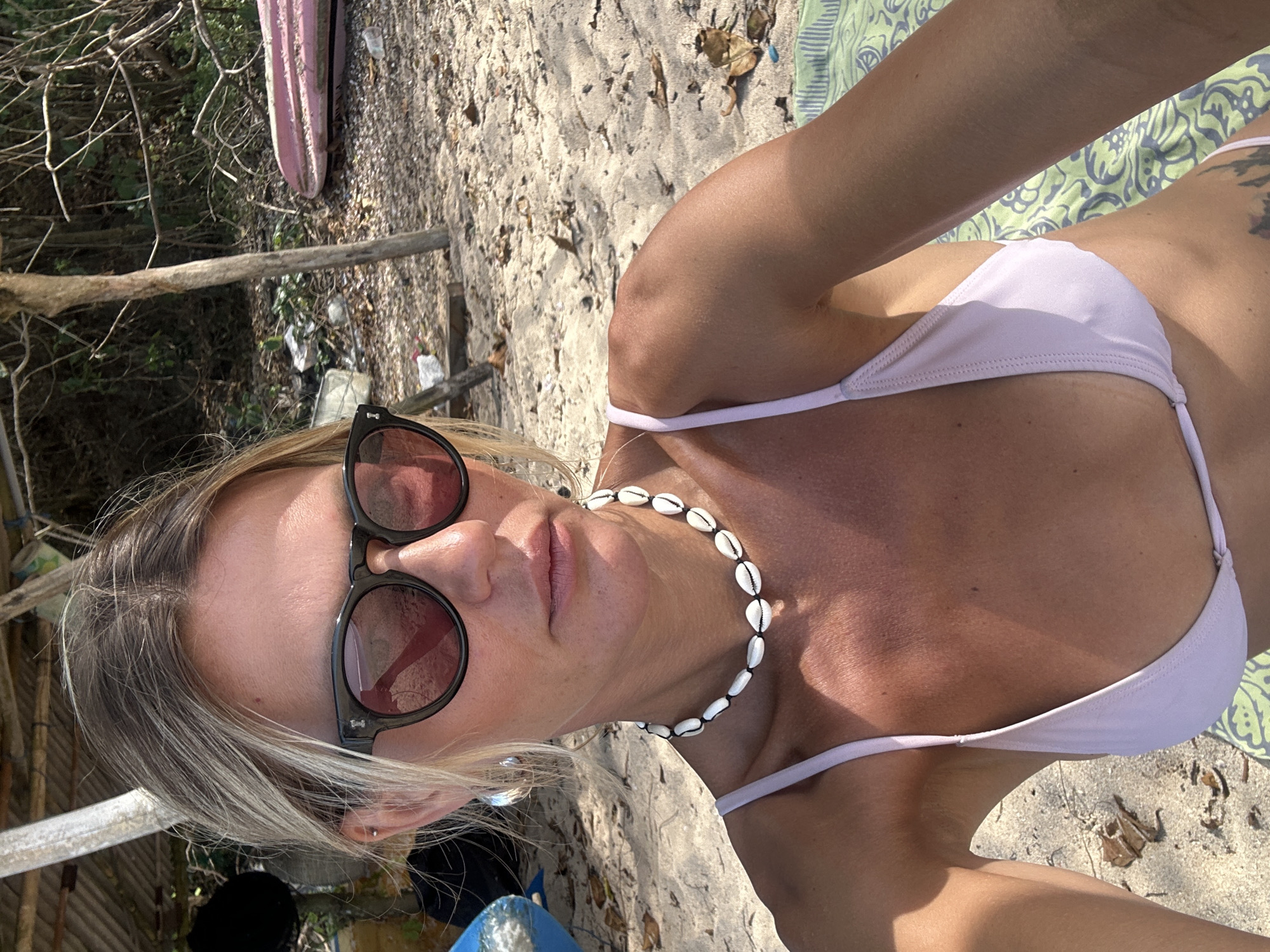 A woman with blonde hair pulled back and wearing sunglasses and a light purple bikini top is pictured on a sandy beach. She has a shell necklace around her neck. Her left arm is resting on a green and white patterned towel. In the background, there is vegetation, a rustic wooden structure, and two pink objects that appear to be surfboards or kayaks. The scene suggests a sunny day on a beach in Kuta Selatan, Indonesia.