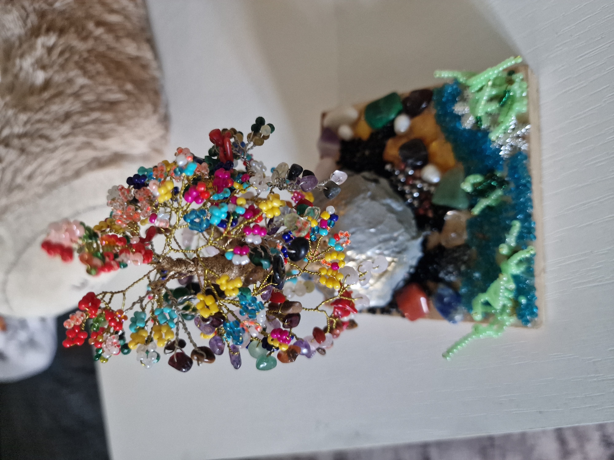 The image shows a close-up, eye-level view of a decorative beaded tree positioned on a white wooden surface. The tree, crafted with multi-colored beads resembling flowers and leaves, is mounted on a base adorned with various stones and resin that creates a landscape effect with blue and green elements. Small green plastic figures, resembling dinosaurs or reptiles, are placed on the left side of the base. The background is out of focus, showing a light-colored, textured object. There is no indication of the city of Kursavka, Russia, or any other specific location.