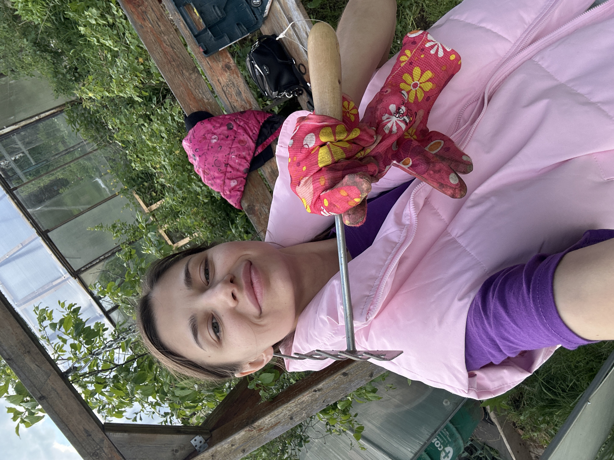 A young woman is outdoors near a greenhouse, possibly in the vicinity of Khokhryaki, Russia. She is wearing a pink jacket and a purple long-sleeved shirt. She is holding a garden cultivator with gardening gloves on her hands. A pink quilted hat and a small tool bag are visible on a wooden structure behind her. There are plants and green vegetation around the greenhouse.