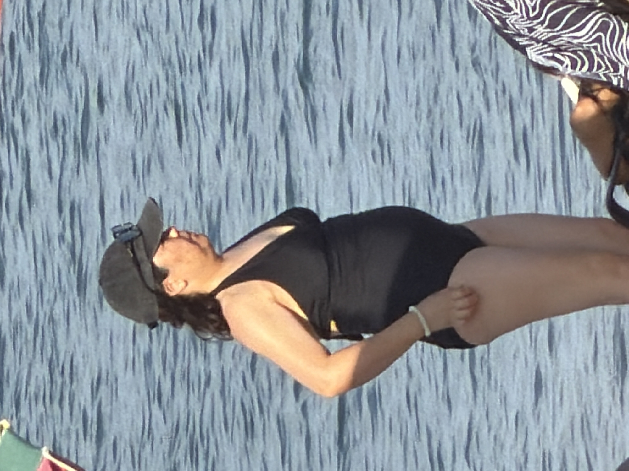 A person wearing a black one-piece swimsuit, a black baseball cap, and sunglasses stands in the water. The person's left arm is bent at the elbow, and they are wearing a light-colored bracelet on their left wrist. The background is the ocean with gentle waves.