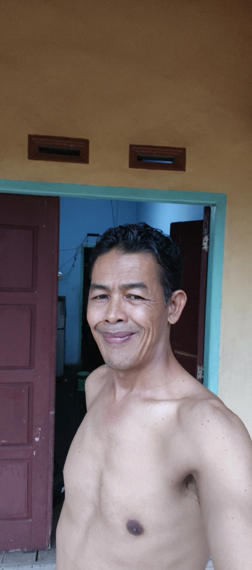 A man is standing shirtless in front of a building with an orange wall and a brown door. He has dark, wavy hair and is smiling. The man's chest and shoulders are visible. In the background, there is a blue door and an interior room. The setting appears to be a residential area.