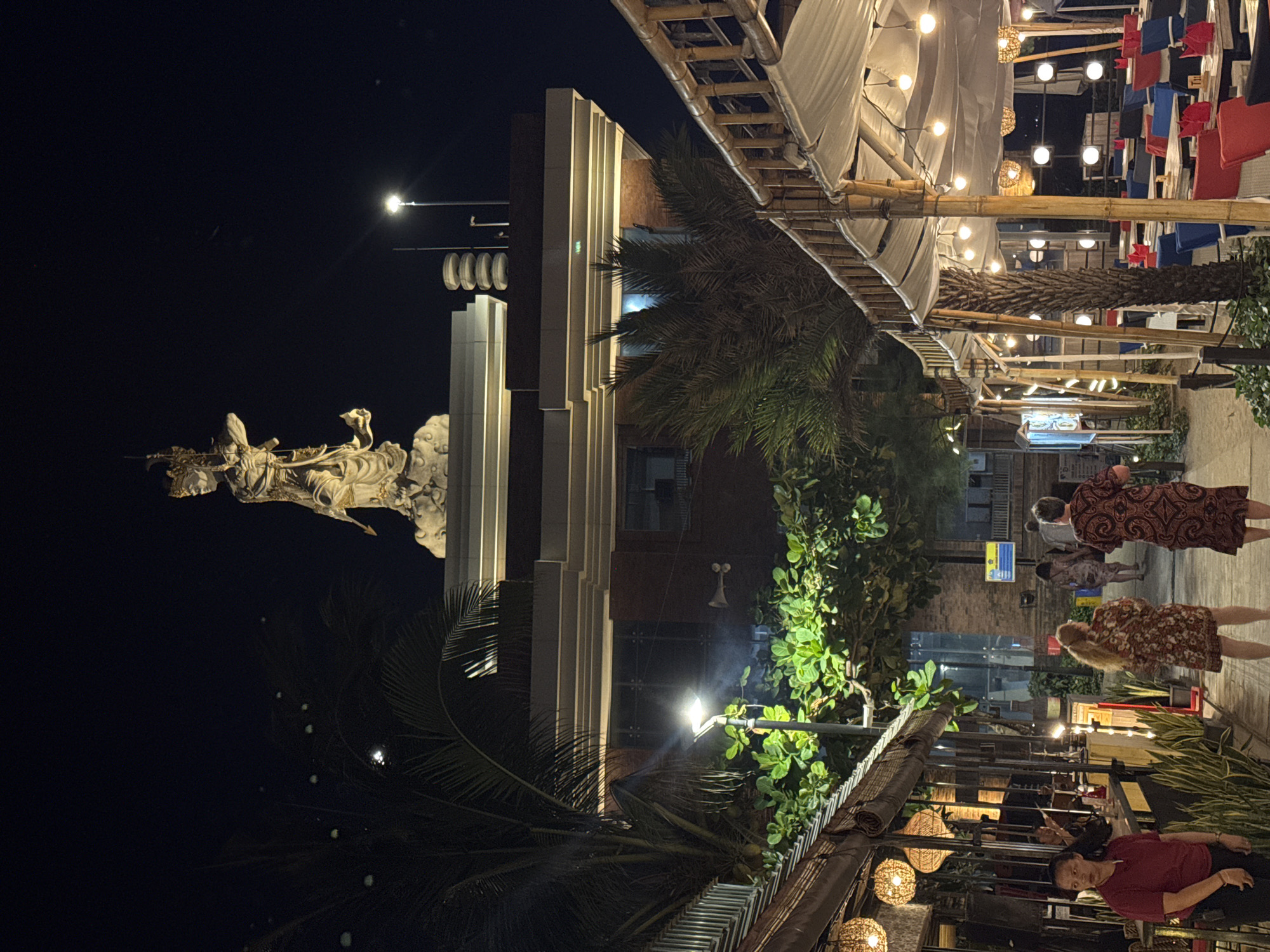 The image depicts an exterior view of a bustling street in Kuta, Indonesia at night. A large, illuminated statue of a deity stands atop a multi-story building in the background. The foreground features an outdoor dining area with tables and chairs under bamboo-framed canopies, decorated with string lights. Several people are visible, including a woman in a red shirt in the foreground, and two women in patterned dresses walking away from the camera. The environment is characterized by palm trees, lush green foliage, and streetlights. The overall atmosphere suggests a vibrant nightlife scene, likely a tourist area given the prominent statue and outdoor dining setups.