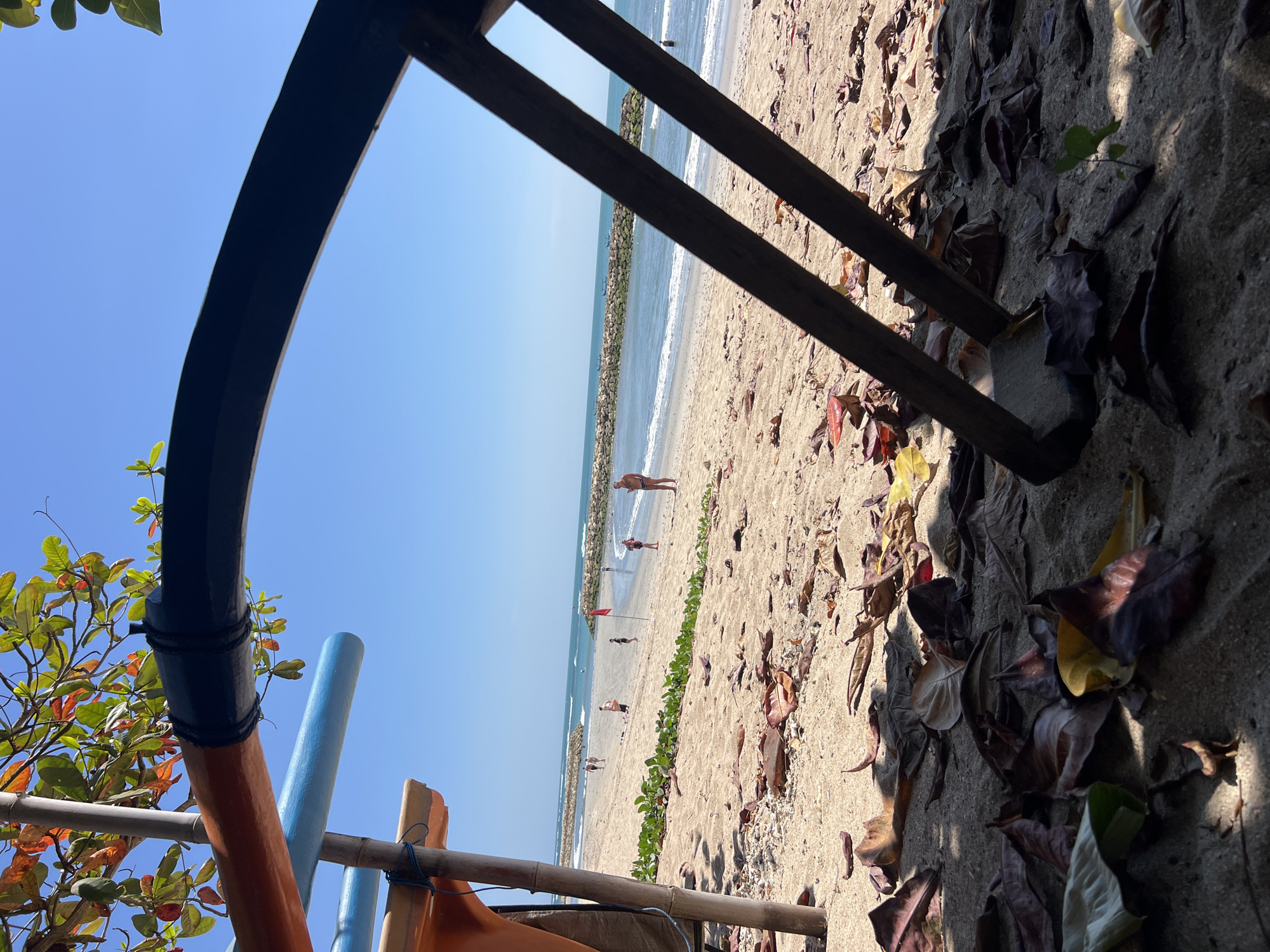The image shows a section of Kuta Beach in Bali, Indonesia, framed by the colorful parts of a boat and some foliage. In the foreground, the sand is covered with fallen leaves, casting shadows. The middle ground features the sandy beach with a few people scattered around, some walking and others near the water. A red flag is planted in the sand. The ocean stretches to the horizon, with gentle waves breaking near a breakwater made of rocks. The sky is a clear, bright blue.