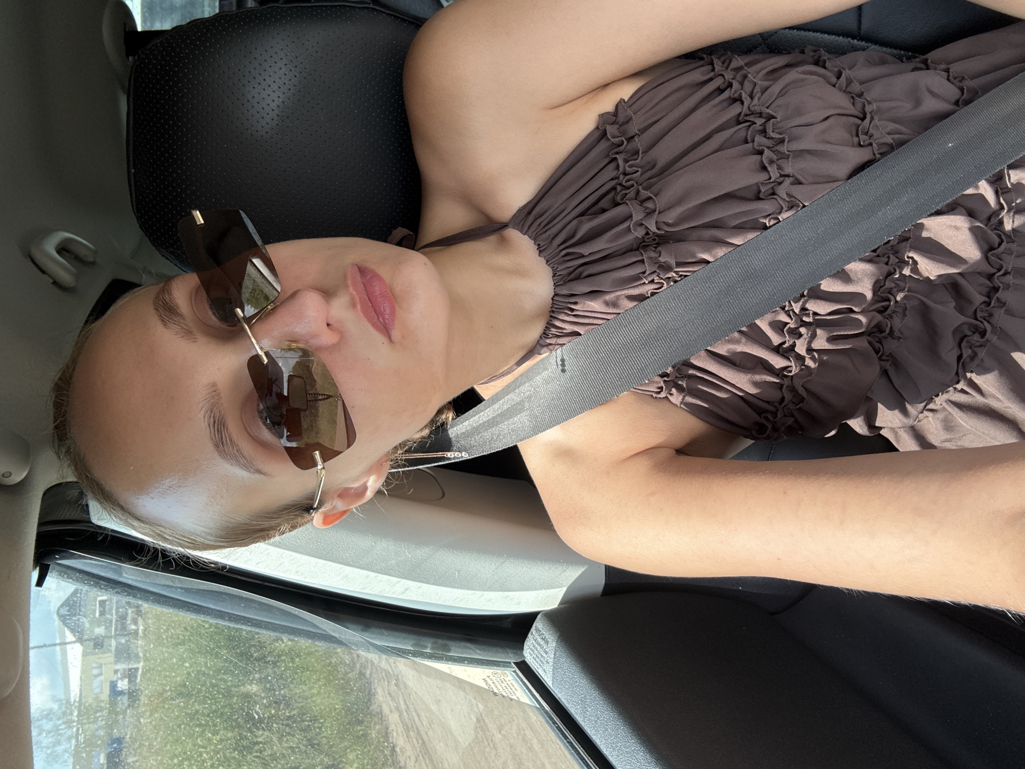A young woman with blonde hair pulled back is seated in the passenger seat of a car, wearing oversized brown sunglasses and a brown ruffled halter dress. She is looking towards the camera. Her seatbelt is fastened across her chest. Through the car window to her left, a sun-drenched landscape with greenery and buildings is visible. The interior of the car, including the black leather headrest and part of the dashboard, is also visible. The image appears to be taken on a sunny day. The approximate location is Pirogovo, Russia.