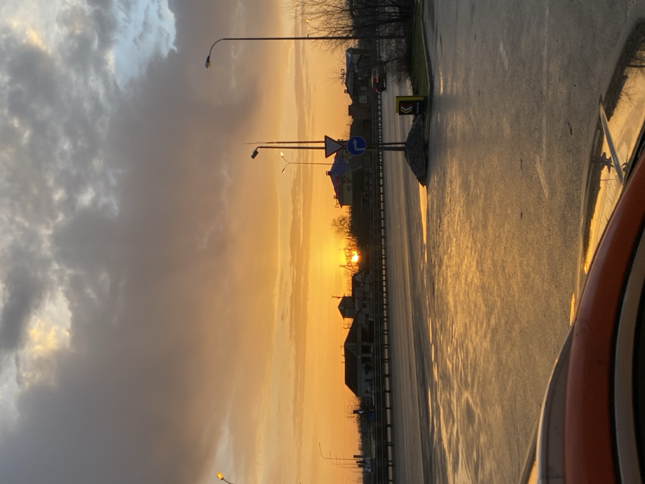 A street scene at sunset, taken from inside a vehicle. A wet, reflective road stretches into the distance, reflecting the warm hues of the setting sun. Houses line the street, silhouetted against the orange and yellow sky. Streetlights and road signs are visible along the road. A storm cloud hangs to the left, contrasting with the bright sky and adding dramatic depth to the scene. Part of the vehicle's interior is visible on the right.