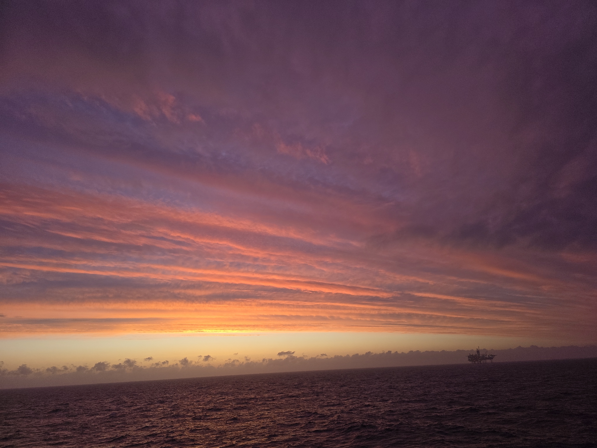 The image depicts a seascape at sunset or sunrise. The sky is dominated by layers of clouds in shades of purple, pink, orange, and yellow. A thin band of orange light is visible on the horizon. The sea is a dark color. In the distance, there appears to be an offshore oil rig. The location is unknown.