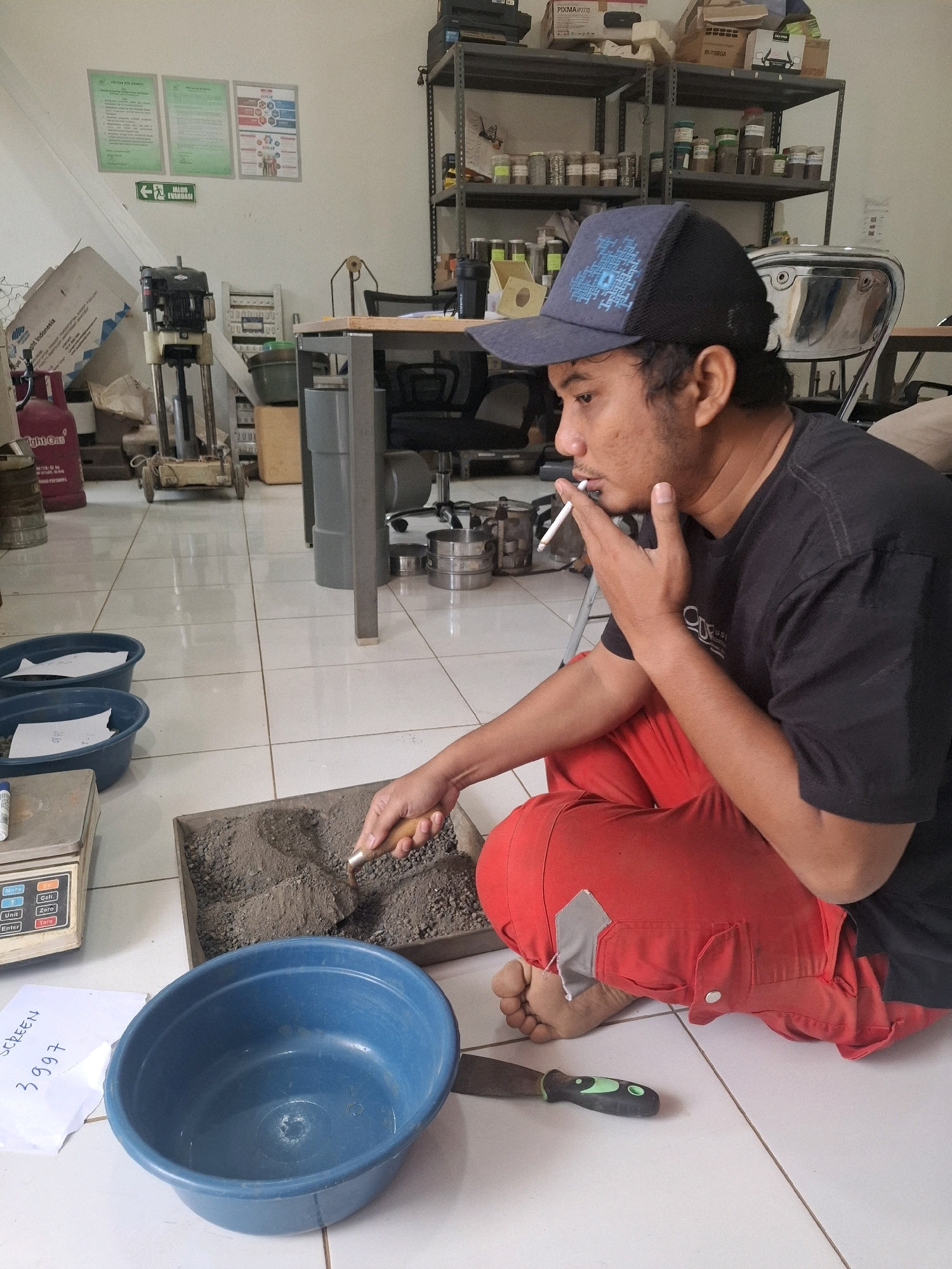 In an indoor laboratory setting, a male individual wearing a black t-shirt and red pants is crouched on a tiled floor. He has a dark blue baseball cap on his head and is smoking a cigarette, holding it between his right index finger and thumb. In front of him, on the floor, is a rectangular tray filled with grey granular material, which he is actively working on with a small trowel. To his left, there are two blue plastic bowls and a digital scale. A piece of white paper with handwritten text is also visible near the scale. The environment appears to be a functional workspace with various laboratory equipment and supplies visible in the background, including shelves stocked with jars and containers, a large metal cylinder, and a gas cylinder. The lighting is even, suggesting overhead illumination. The overall scene depicts a person engaged in a task within a laboratory or workshop.