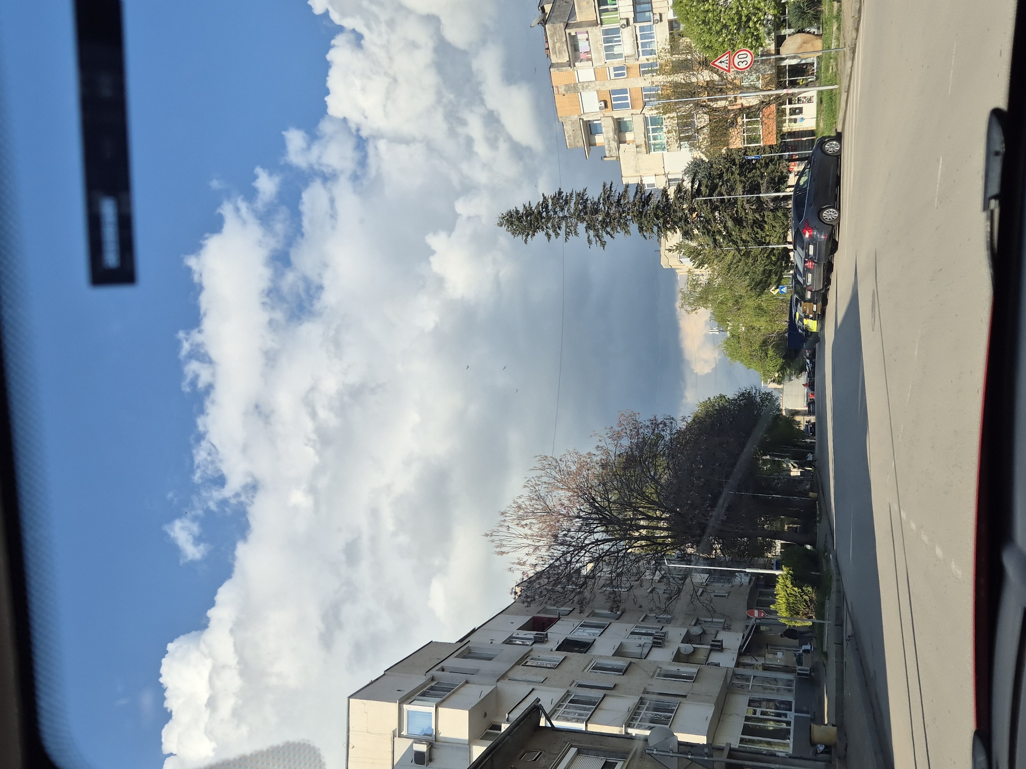 The image presents an urban street scene in Pavlikeni, Bulgaria, captured from the interior of a vehicle, as indicated by parts of the car frame visible on the left and right edges. The street runs from the foreground towards the background, flanked by buildings and trees under a dynamic sky.
On the left side of the street, multi-story residential buildings line the pavement. These structures feature numerous windows, balconies, and exterior air conditioning units, typical of mid-20th century Eastern European architecture. Various trees are interspersed along the street; a prominent tall, slender evergreen stands in the middle distance, while another tree with delicate, reddish-brown foliage is visible further down.
The sky above is a significant element, showcasing patches of bright blue amidst large, puffy white cumulus clouds. In the upper center, a darker, greyish cloud formation suggests mixed weather conditions. Several tiny, dark specks, possibly birds or insects, are visible against the sky. Thin overhead wires traverse the scene.
Along the right side of the asphalt road, several cars are parked, including a dark-colored sedan in the mid-ground. Street signs are visible, including a red-bordered circular sign displaying "30," indicating a speed limit, and another red circular sign with a white horizontal bar, signifying "no entry" or "do not enter."
The scene is set during daytime, with clear sunlight and visible shadows indicating either morning or early afternoon. There are no prominent human figures or specific activities taking place, portraying a calm, everyday street environment.