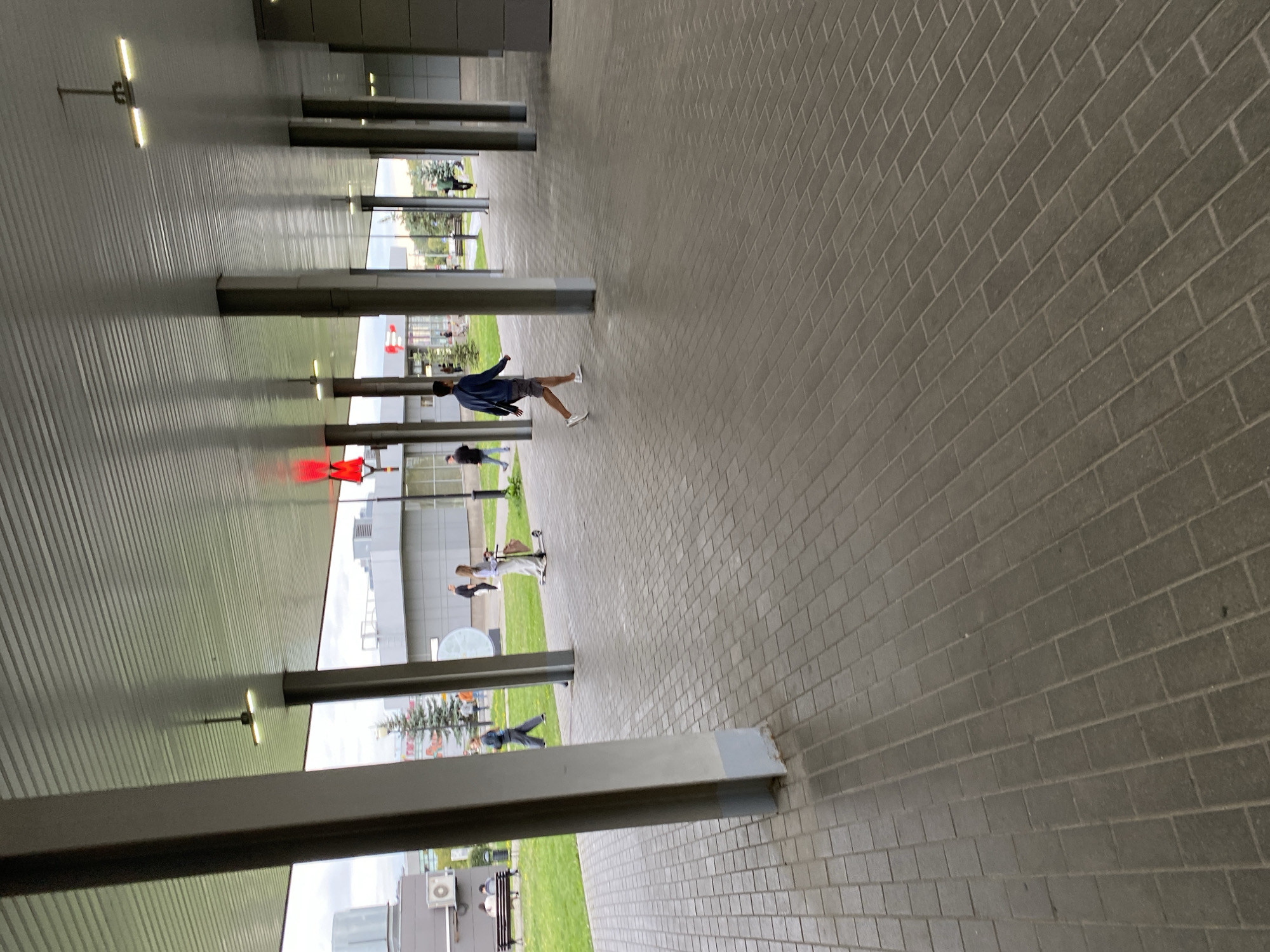 The image shows an outdoor covered walkway with a tiled floor. Several people are visible walking and one person is on a scooter in the background on a grassy area. The walkway is supported by large concrete pillars. The ceiling of the walkway is a ribbed metal structure with industrial lighting fixtures. There are also red decorative elements visible on the ceiling in the distance. The environment appears to be a modern urban setting, likely a public space or the entrance to a building. Given the aesthetic and building style, this scene could be located at the Moscow Metro Station VDNKh, specifically along the covered pedestrian transition from the metro exit to the main exhibition center.