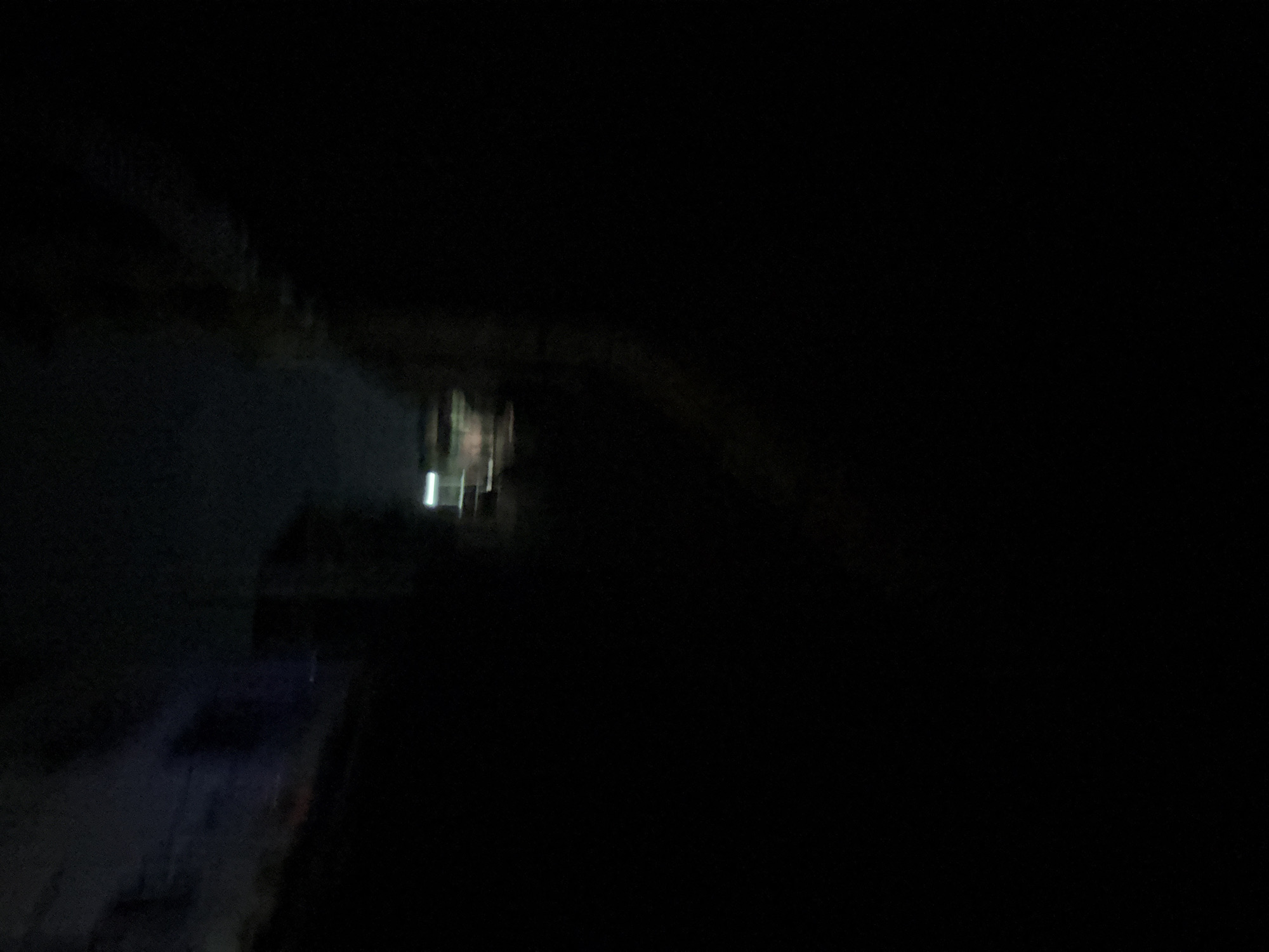 This is a nighttime, out-of-focus, low-light photograph of a street scene in Onitsha, Nigeria. The image is predominantly dark, with minimal discernible details. In the upper central portion of the image, there are faint horizontal streaks of light, likely from vehicle headlights or streetlights, indicating a road or pathway. To the right of these lights, a blurred, dark shape suggests the presence of a building or structure. The overall environment appears to be an urban setting at night, characterized by darkness and sparse artificial illumination.