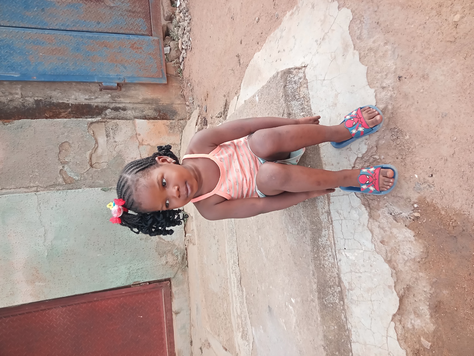 A young, dark-skinned girl sits on a concrete surface in what appears to be an urban environment. She wears a peach and white striped tank top and light-colored shorts. Her hair is styled in braids with a colorful flower hair accessory. She is looking directly at the camera. She is wearing blue sandals with a red and blue cartoon character on the toes. The background consists of a rusty metal door, a weathered wall, and a dirt and concrete ground. The scene is likely located in Sabon Tasha, Nigeria, based on the prompt.