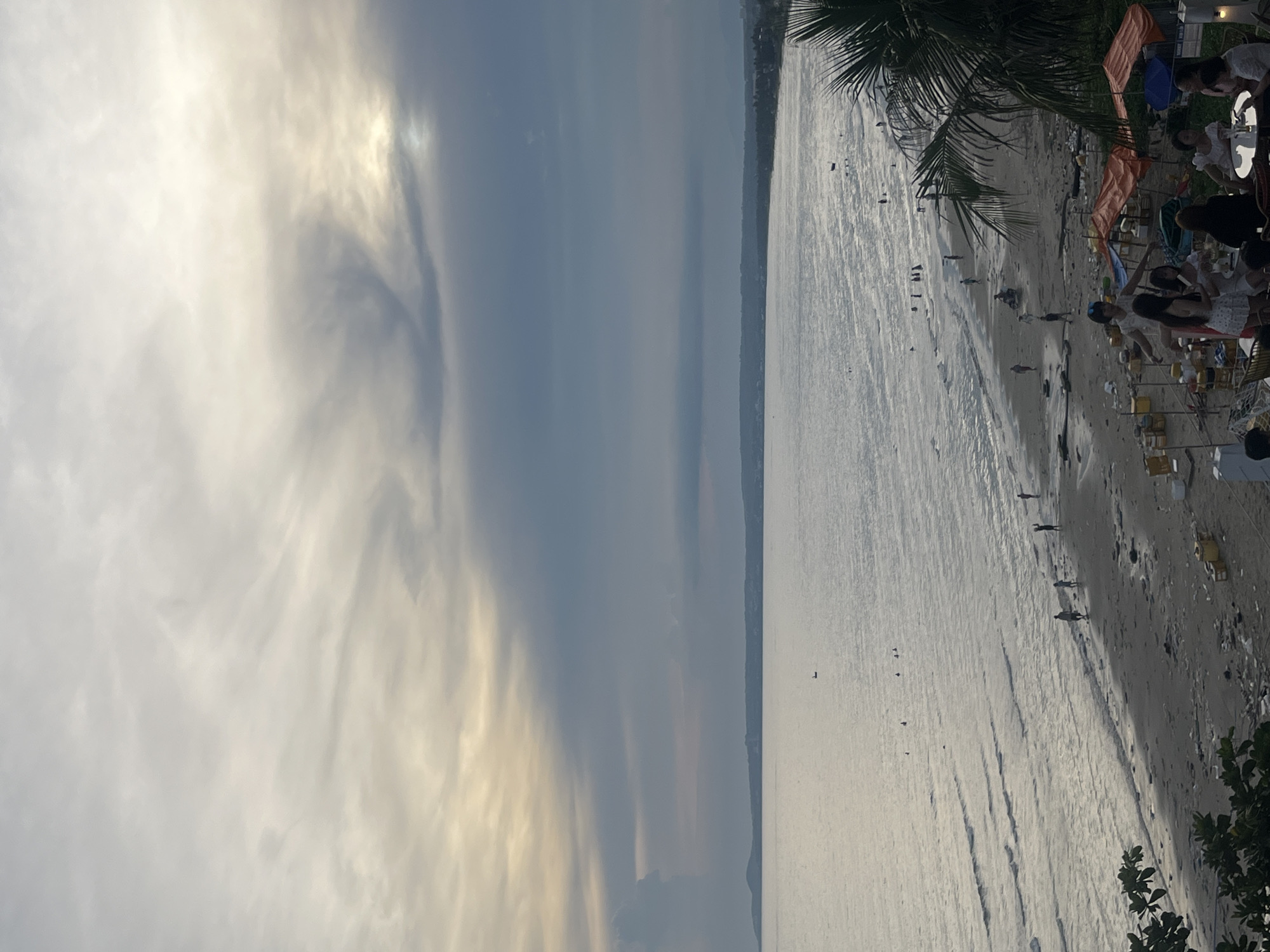 The image depicts a beach scene in Mũi Né, Vietnam, likely during the late afternoon or early evening, given the soft, diffused light and the long shadows. The sky is filled with dramatic, wispy clouds, some illuminated by the sun which is partially obscured, creating a gentle, atmospheric glow.
The scene is divided into two distinct halves: the sky and the sea. The left side of the image is dominated by the expansive, cloud-filled sky, with areas of bright light and darker, more dramatic cloud formations. The right side features the ocean, with small waves gently rolling towards the shore. The water appears silvery and reflective under the fading sunlight.
The shoreline is a sandy beach dotted with small figures of people walking or standing. Some appear to be enjoying the water's edge, while others are strolling along the sand. There are also a few scattered objects on the beach, possibly debris or small structures. Further along the beach, in the upper right portion of the image, there are several makeshift shelters or umbrellas and tables, suggesting a small gathering of people at a beachside establishment. A few individuals are seated at these tables. A portion of a palm tree's fronds is visible in the top right corner, further confirming the tropical beach setting.
The overall mood is serene and peaceful, with a sense of quiet enjoyment of the beach environment. The weather appears to be fair, with no indication of rain, but the cloud cover suggests a transition in the day. There is no visible text in the image.