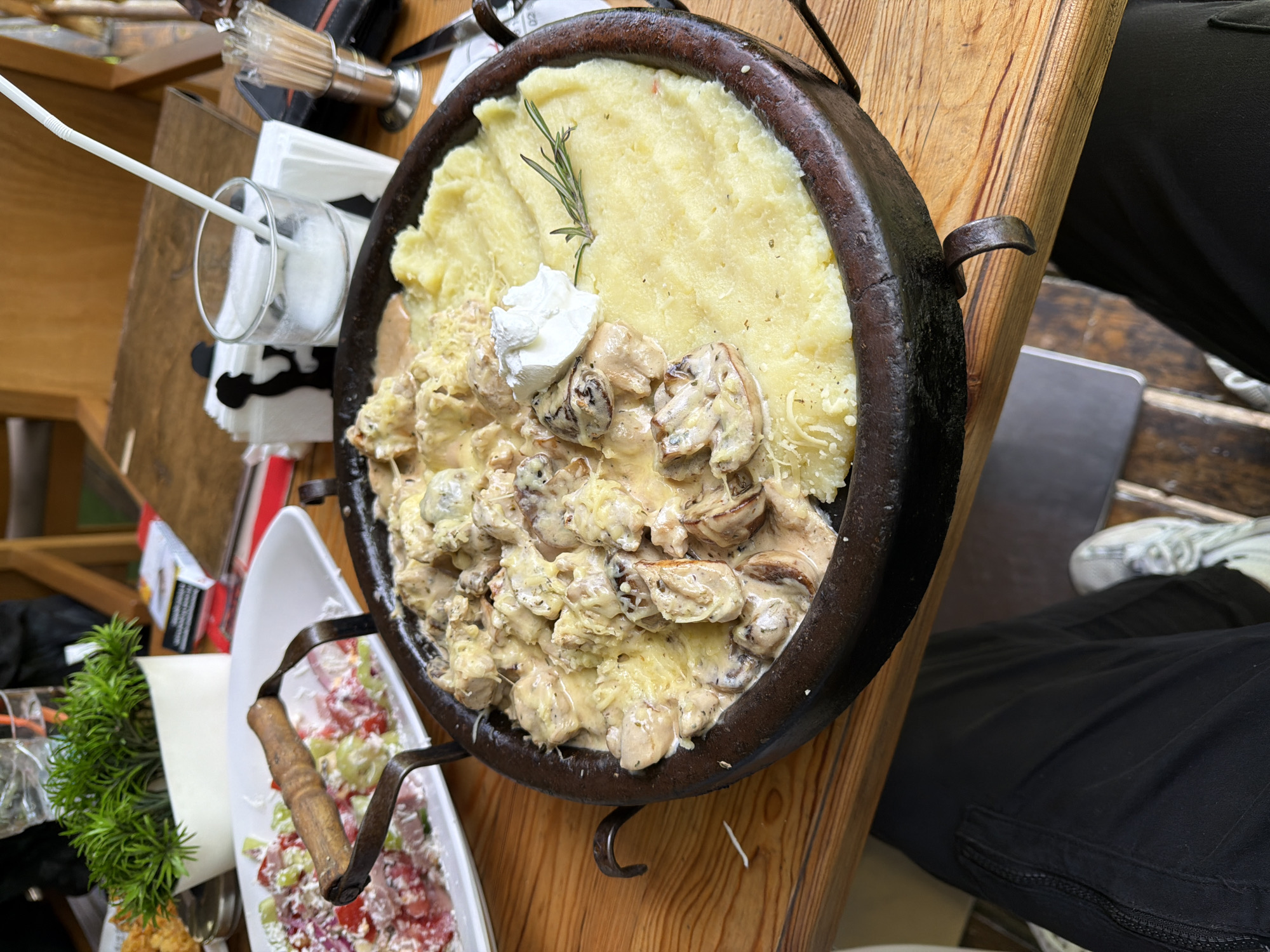 The image shows a large, dark metal pan filled with food, likely served on a wooden table in a restaurant. The pan contains a significant portion of mashed potatoes, with a dollop of cream and a sprig of rosemary on top. Adjacent to the potatoes is a creamy mushroom dish with what appears to be shredded cheese. A glass with a white straw is nearby, along with a small plate containing a salad with red and white ingredients. Part of a potted plant can also be seen. The scene appears to be indoors, probably within a restaurant. The image was taken in Blagoevgrad, Bulgaria. The image also shows parts of a person: their black trousers and white sneakers.
