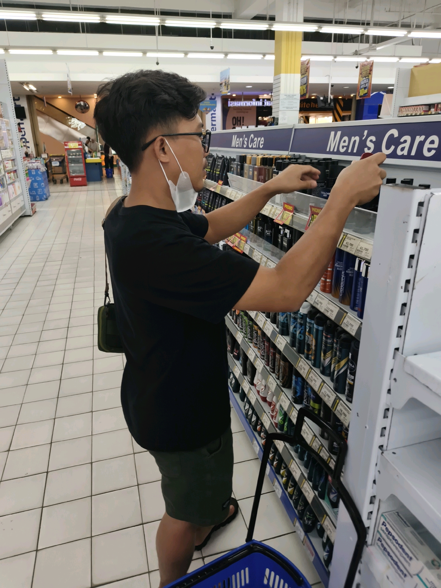 A young man wearing glasses, a black t-shirt, and green shorts is standing in the men's care aisle of a brightly lit supermarket in Ketapang, Indonesia. He has a white face mask hanging below his chin. His left hand is reaching out towards a shelf stocked with various grooming products, including spray cans. His right hand is near the top of the shelf, also appearing to be browsing. A dark blue shopping basket is visible at the bottom of the frame, with a black shopping cart handle extending upwards. The floor is tiled in a white square pattern, and fluorescent lights illuminate the scene from above. In the background, there are more aisles of merchandise, promotional signs, and other shoppers.