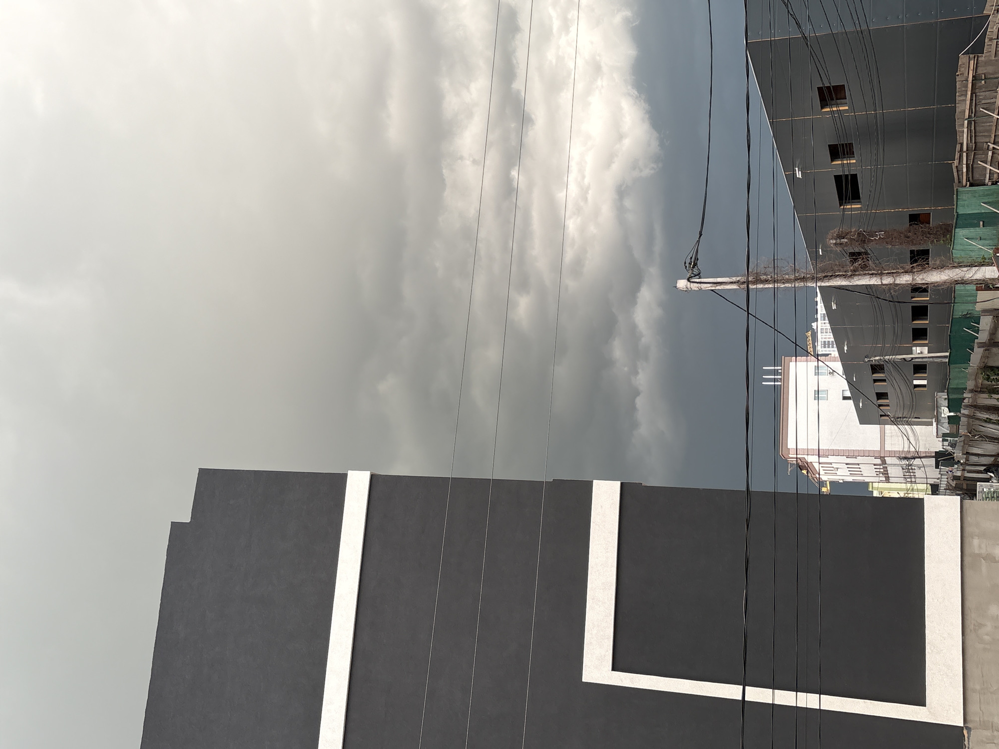 The image shows a cityscape in Hatne, Ukraine, taken during an overcast day with a gray sky and heavy clouds. In the foreground, there is a dark gray building with white rectangular accents. Power lines run vertically across the frame, cutting through the sky and buildings. In the middle ground, there is a taller, black building with reflective windows, partially obscured by construction materials and green netting. Adjacent to this is a white building with multiple floors. The buildings are arranged close to each other, creating a dense urban environment. The focus appears to be on the juxtaposition of modern architecture against the backdrop of a stormy sky. There are no people visible in the image.