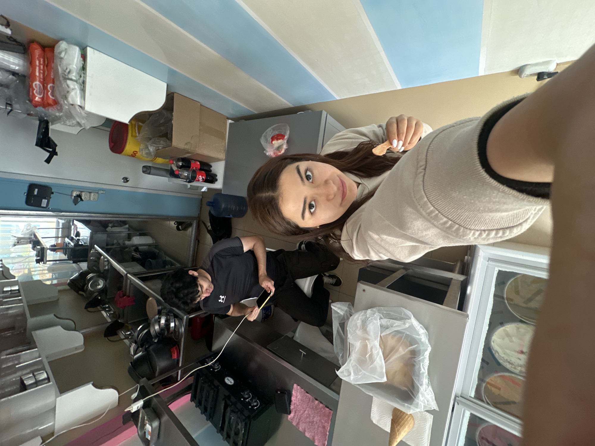 The scene depicts two individuals inside what appears to be an ice cream shop in Puebla, Mexico, based on the environment and ambiance of the shop. A young woman, facing the camera, holds a small wooden spoon or taste tester, seemingly having sampled the ice cream. She has long brown hair, fair skin, and is wearing a beige sweatshirt. In the background, a young man wearing a black t-shirt is focused on his phone, standing near a stainless steel counter with what looks like a stove. The environment includes ice cream display cases, kitchen equipment, and storage shelves. The ceiling has alternating stripes of light blue and white. There are ingredients and supplies stored on the shelves. The shop appears to be in the midst of preparation or operation.