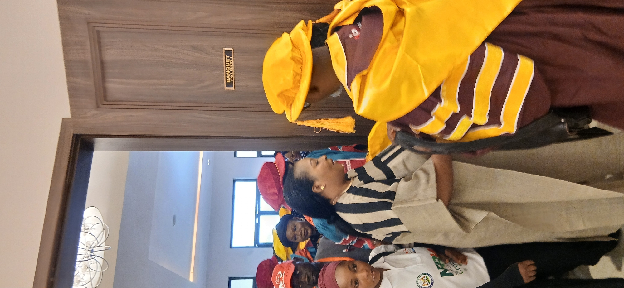 The image captures an indoor scene at what appears to be an academic or celebratory event. In the foreground, a person is seen wearing full academic regalia, consisting of a maroon gown, a bright yellow hood, and a yellow academic cap with a tassel. They stand near a wooden door, facing into a hall.
To the right, a woman with dark hair and a gold earring is smiling and looking towards the person in regalia. She is dressed in a black and white striped short-sleeved top and a light, flowing skirt. Behind her, several other people are gathered inside the hall. Among them, some individuals are wearing red caps, another wears a yellow cap, and one person is in a white t-shirt with a partially visible logo.
The setting appears to be a modern banquet hall, characterized by sleek design elements like recessed ceiling lighting and large windows that allow natural light into the space, indicating it is daytime. A significant detail is a small rectangular plaque on the wooden door, which clearly displays the text "BANQUET HALL ENTRY". The door itself features light-toned wood with decorative paneling. The overall mood conveyed is one of positive engagement and gathering, consistent with a formal event or ceremony.
