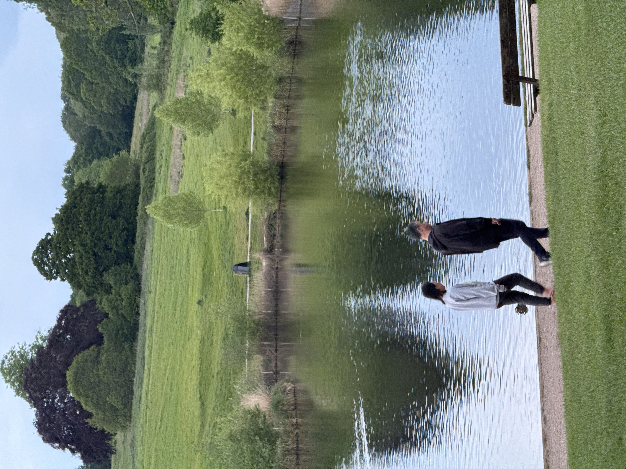 Two people are standing near a lake on a grassy area. One person is wearing a light-colored top and dark-colored pants, and is closer to the lake edge. The other person is wearing a dark-colored top and dark-colored pants. The lake reflects the sky and the surrounding grassy area. Trees are visible along the edge of the grassy area, and a bench is positioned on the edge of the lake. The location is in London, United Kingdom.