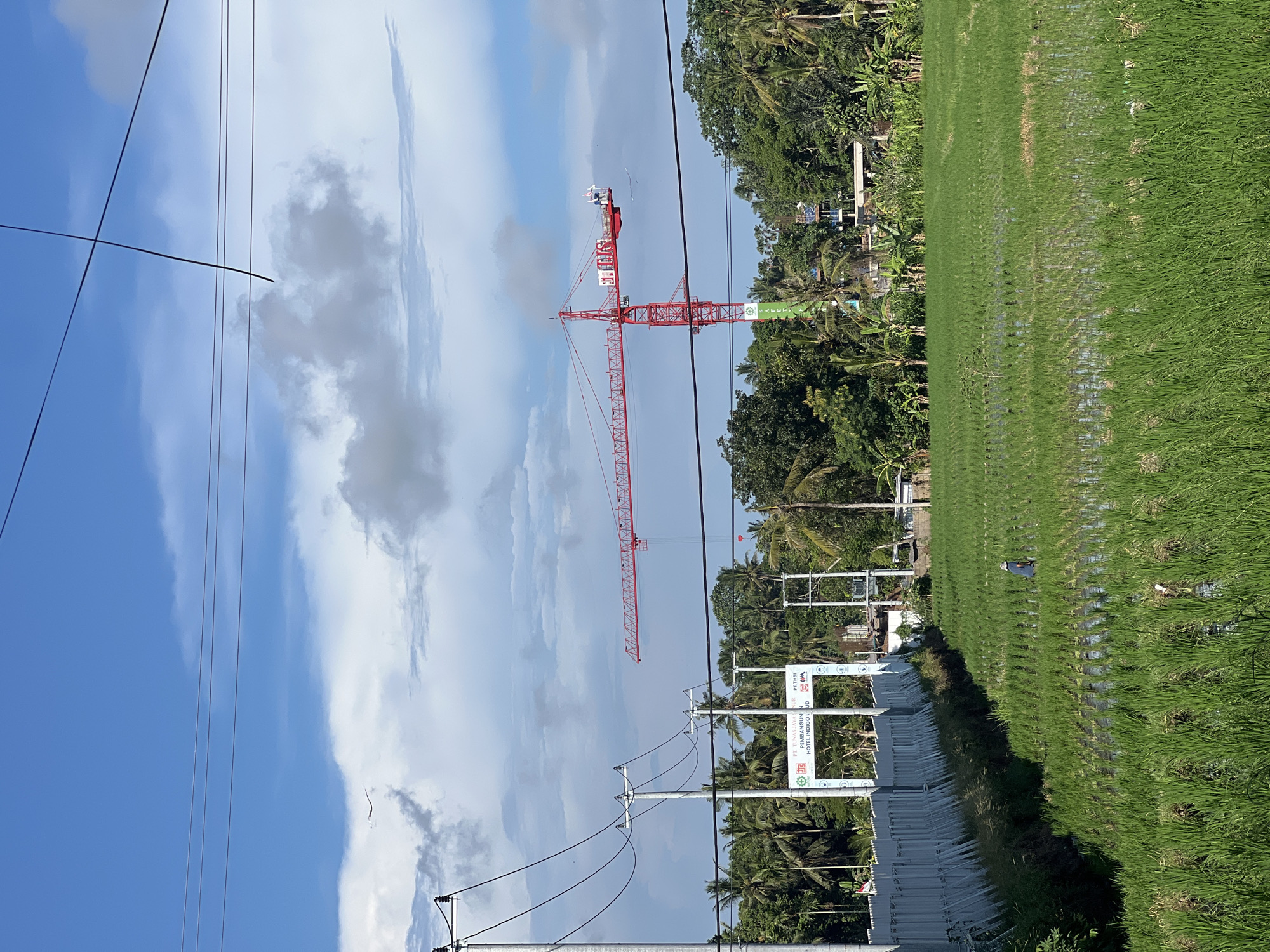 The image captures a scene in Sukawati, Indonesia, depicting a lush green rice paddy field adjacent to a construction site. A tall red construction crane dominates the skyline against a backdrop of a partly cloudy sky. Overhead power lines traverse the scene, adding a network of linear elements. A series of construction signs stand along the edge of the field, possibly indicating the project being undertaken. In the distance, dense tropical vegetation and palm trees line the horizon, suggesting a location in the countryside or outskirts of the city. A person is seen in the rice paddy field.