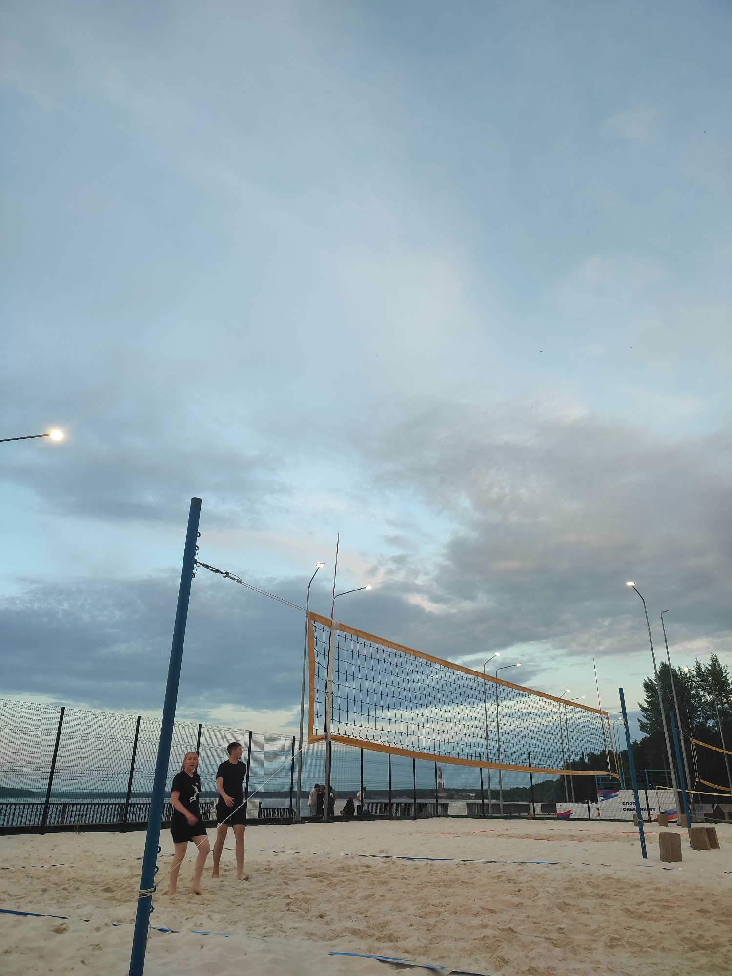 This image captures a beach volleyball court at dusk. Two individuals, a woman and a man, both dressed in black athletic wear, are positioned on the sand in front of a volleyball net. They appear to be playing or preparing to play. The court is sandy, with blue boundary lines visible. A volleyball net, supported by blue poles, stretches across the court. In the background, a body of water is visible beyond a fence, with trees and a distant structure on the horizon. Several streetlights are positioned around the court, indicating it is used in the evening. The sky is overcast with dramatic clouds, suggesting either approaching rain or the setting of the sun. The overall scene is calm and recreational.