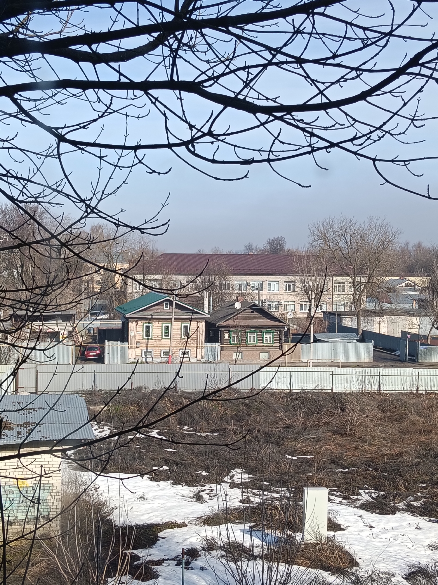 The image depicts a residential scene in Murom, Russia, viewed through bare tree branches in early spring, indicated by remaining patches of snow on the ground. Two traditional wooden houses, one with a green roof and light-colored siding, the other darker with green trim, are situated behind a corrugated metal fence. A red car is visible in a yard behind the fence, next to the green roofed home. In the background, a large, multi-story brick building with a brown roof can be seen. The foreground features a mix of snow, dead grass, and small shrubs. A small brick building with graffiti is visible to the left. A white rectangular object is present at the bottom right. The sky is pale blue.