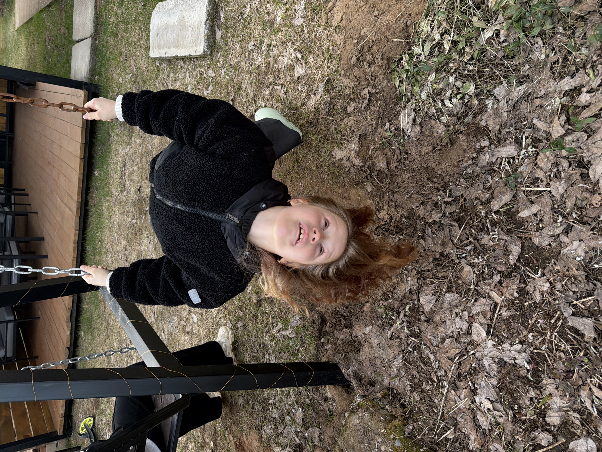 A young child with fair skin and light brown, wavy hair is captured in an outdoor setting, likely in Markovo, Russia. The child is positioned as if looking upwards, with a wide, joyful smile revealing their teeth. They are wearing a black, fuzzy fleece jacket, dark pants, and distinct footwear: a light green boot or shoe on their left foot and a white sneaker on their right. The jacket features the text "FOREVER" on the left chest area.
The child holds onto rusty metal chains with both hands, suggesting they are part of a swing or an outdoor play structure. These chains are attached to a black metal frame, around which thin, golden string lights are decoratively wrapped.
The ground beneath and around the child is a mix of bare, light brown dirt, an abundance of dry, brown leaves, and sparse patches of green grass and small weeds, indicating a natural, possibly less maintained, outdoor area, perhaps in early spring or late autumn. A wooden deck is visible to the left, and concrete blocks are scattered on the ground further back. A small part of a yellow/green wheeled toy is also visible near the black frame. The overall lighting is bright and diffuse, typical of daytime. The child's happy expression conveys a sense of enjoyment and play.