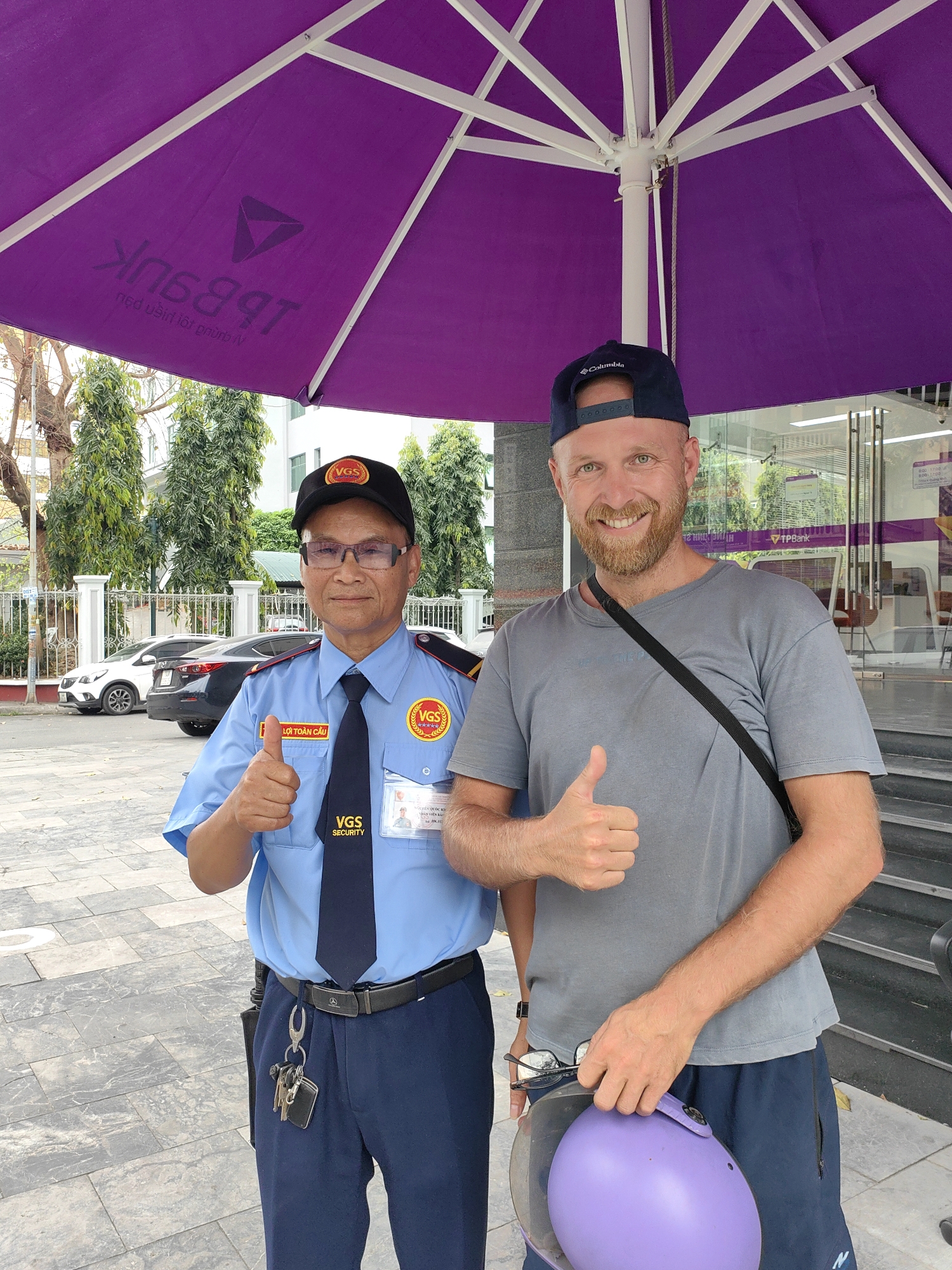 Two men stand under a purple umbrella outside a building. The man on the left wears a blue uniform with "VGS Security" patches, a black tie, and a "VGS" hat. He has glasses and gives a thumbs-up. The man on the right wears a gray shirt, a dark baseball cap, and a black strap across his body. He also gives a thumbs-up and holds a purple helmet in his left hand. The building has a "TPBank" logo. The man on the left is likely located at Hong Gai Ward, Vietnam. In the background are trees, a fence, and parked cars.