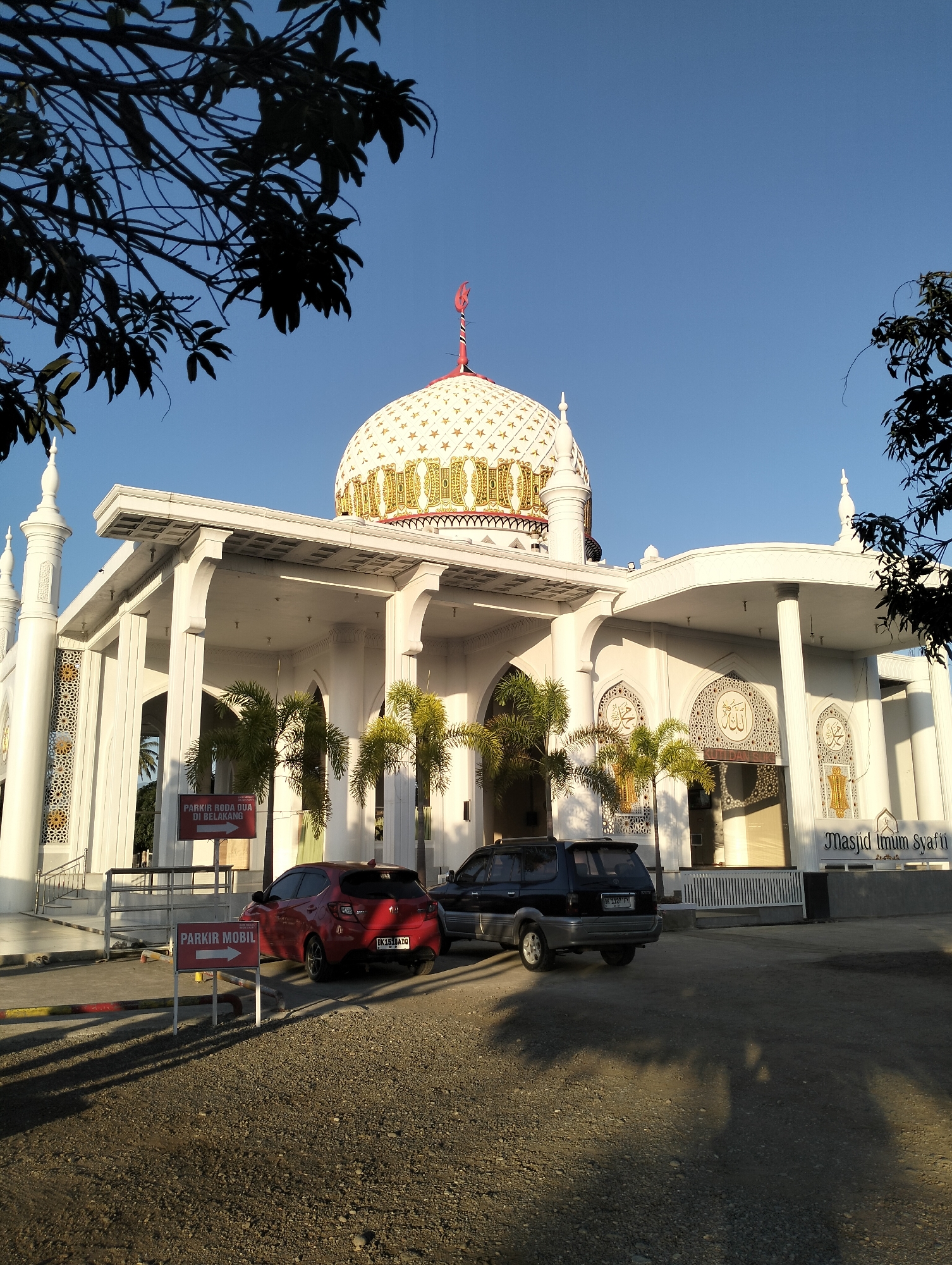 The image shows a bright, sunny day in Blangmetimu, Indonesia, with a clear blue sky. A prominent white mosque with a decorative dome and minarets stands in the center. The mosque features intricate white latticework and ornate gold accents on its dome and facade. In front of the mosque, there are two signs, one indicating "PARKIR MOBIL" (Car Parking) and another pointing to "PARKIR RODA DUA DI BELAKANG" (Two-Wheeler Parking in the Back). Two cars are parked in the foreground: a red hatchback and a dark SUV. Palm trees are scattered around the mosque, and tree branches with dark green leaves frame the top of the image. The ground is a mix of gravel and pavement, with long shadows cast by the trees and the mosque. The overall scene depicts a well-maintained religious site and its surrounding parking area.