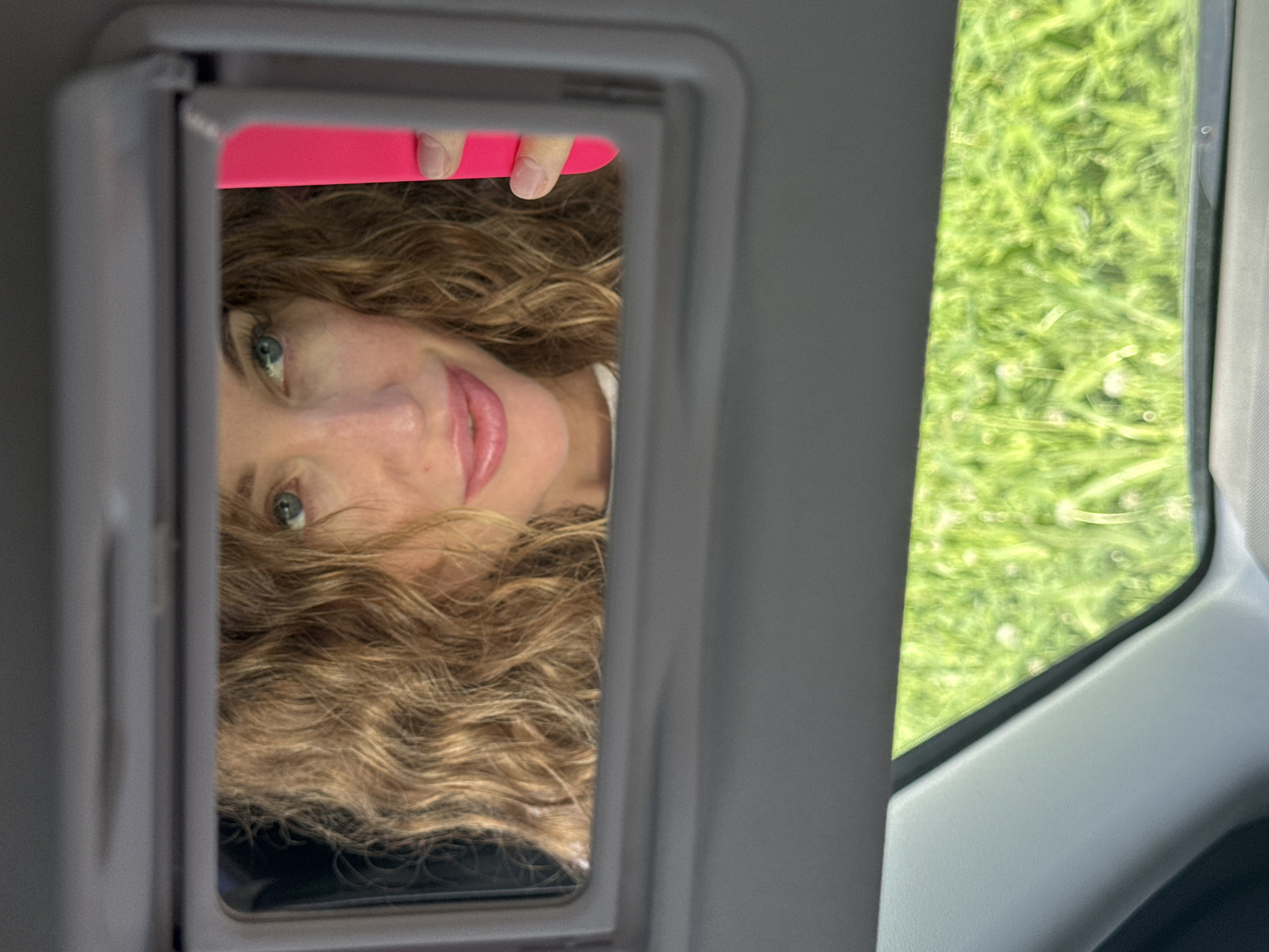 A woman's face is reflected in a car's visor mirror, occupying the left side of the vertically oriented image. Her head is angled, with her long, wavy, light brown hair framing her face. She has light-colored eyes, a subtle smile, and naturally pink lips. Her gaze is directed slightly upwards and towards the viewer. A hand with neatly manicured nails is visible at the top edge of the mirror's reflection, holding a vibrant hot pink object, possibly a smartphone, which is partially obscured.
To the right of the mirror, outside the car window, is a blurred expanse of bright green, indicating foliage like grass or trees. The blur suggests either movement or a soft focus on the background. The car's interior trim, in shades of gray and beige, frames both the mirror and the window. The scene depicts a casual moment during daylight, likely while the car is in motion or parked in a natural environment. The woman appears to be looking at her reflection, possibly taking a photo. No text is visible. The location context is Chaikovsky, Russia.