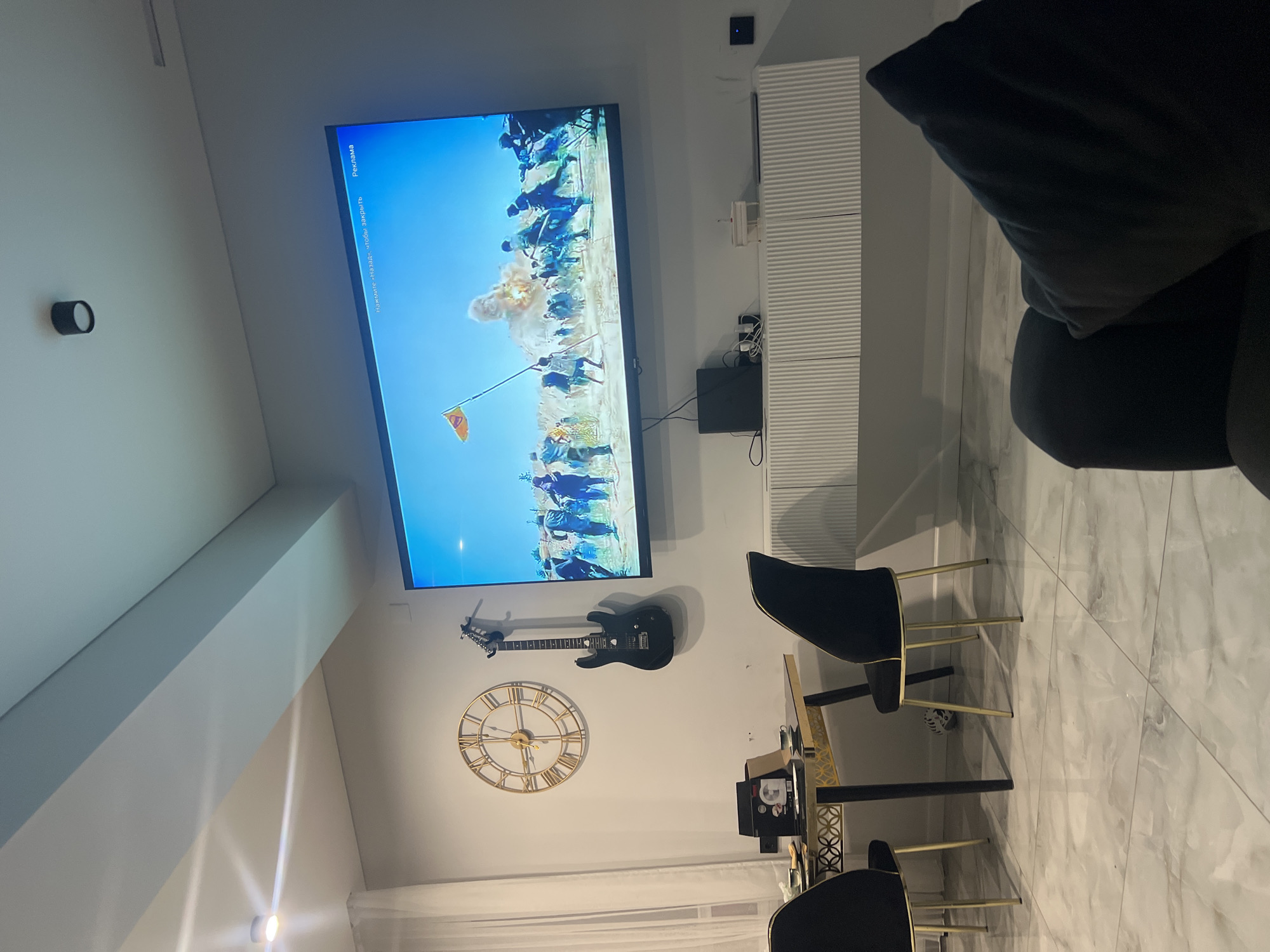 This media file depicts a living room with a large television displaying a scene that appears to be historical or fictional, possibly a battle or a reenactment. The screen shows numerous figures in what looks like a procession or a conflict, with some figures carrying a flag. There is an explosion or smoke on the screen. The room has a modern aesthetic with a white wall and a large, sleek television mounted on it.
Below the television, a black electric guitar is mounted on the wall, adding a decorative element. To the left of the guitar is a large, round gold clock with Roman numerals, indicating the time. In the foreground, a modern dining set with black chairs and a gold-accented table is visible. The floor is tiled with a marble-like pattern. The lighting suggests it could be daytime or evening, with artificial lights illuminating the space.
Visible text on the TV screen reads "Реклама" and "Нажмите «Назад», чтобы закрыть," which translates to "Advertisement" and "Press 'Back' to close," indicating that the content on the screen is an advertisement. The overall scene conveys a sense of a well-decorated, contemporary living space.