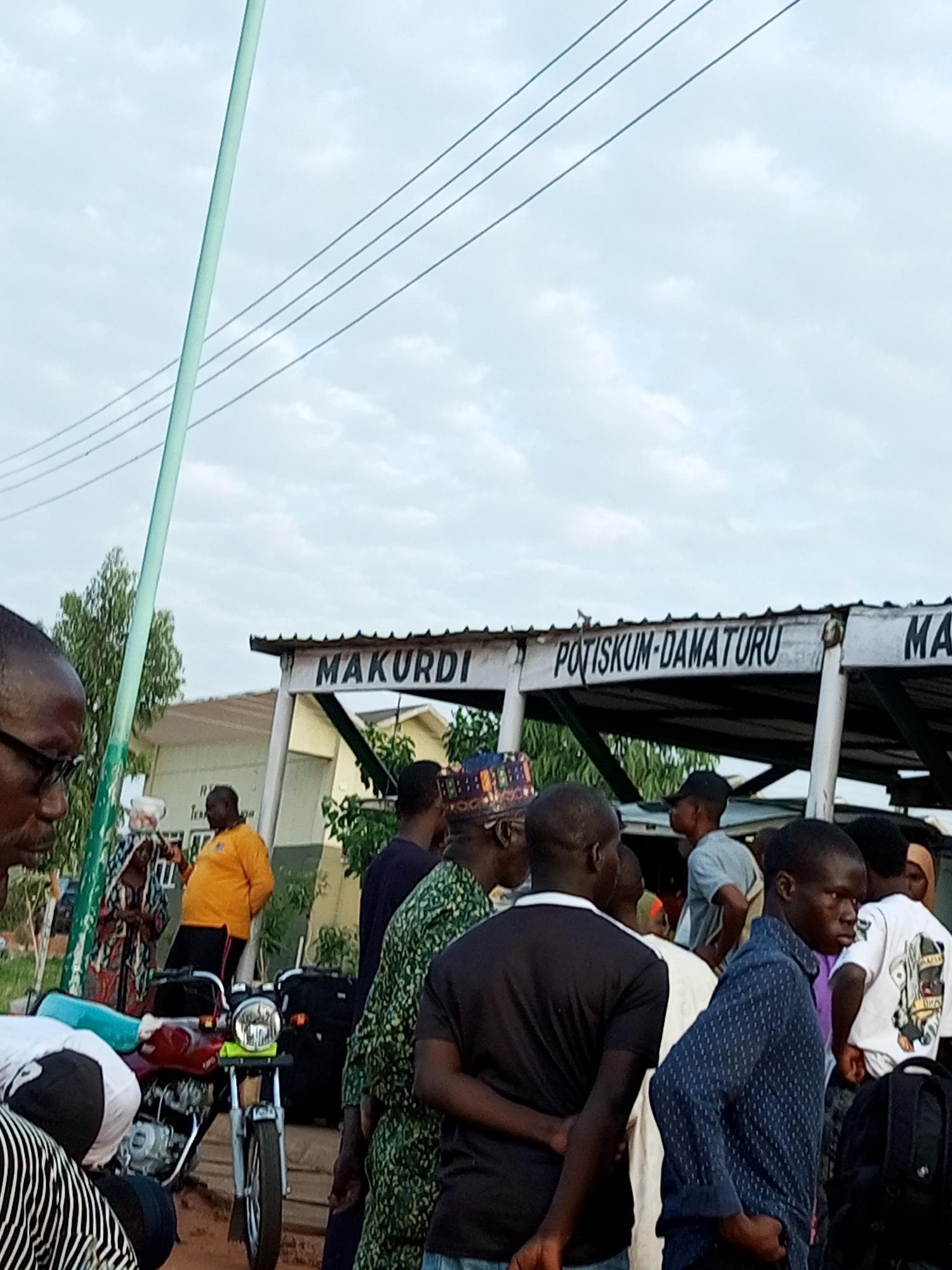 The image depicts a street scene, likely a bus station or transport hub, in the city of Gombe, Nigeria. In the foreground, several men are gathered, some looking away from the camera. A motorcycle is parked in the middle ground, with luggage placed beside it. In the background, a building with signage that reads "MAKURDI POTISKUM-DAMATURU" is visible. The sky is overcast, and power lines are strung across the top of the frame. The overall atmosphere suggests a typical day at a busy transportation point.