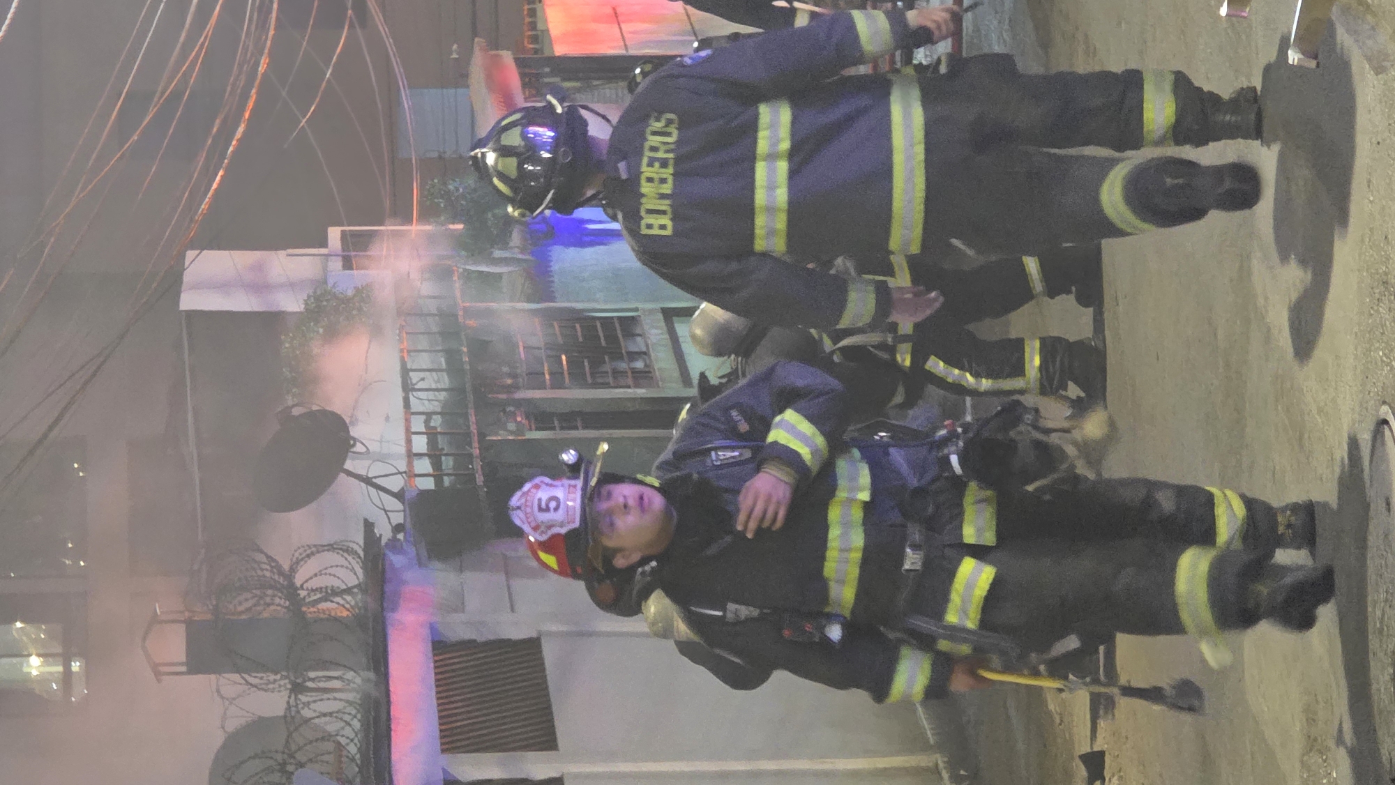 The image depicts a night scene of a fire in an urban environment, with two firefighters in the foreground. Both are wearing full protective gear, including helmets with lights and black uniforms with yellow reflective stripes. "BOMBEROS" is visible on the back of one firefighter's uniform. The second firefighter's helmet has the number "5" on it. The firefighters are standing on a paved surface, possibly a street, near a manhole cover. In the background, there is a building with smoke emanating from it. The building has features like barbed wire and a ladder. Overhead, there are visible power lines and cables. The overall scene suggests an active firefighting operation in progress.