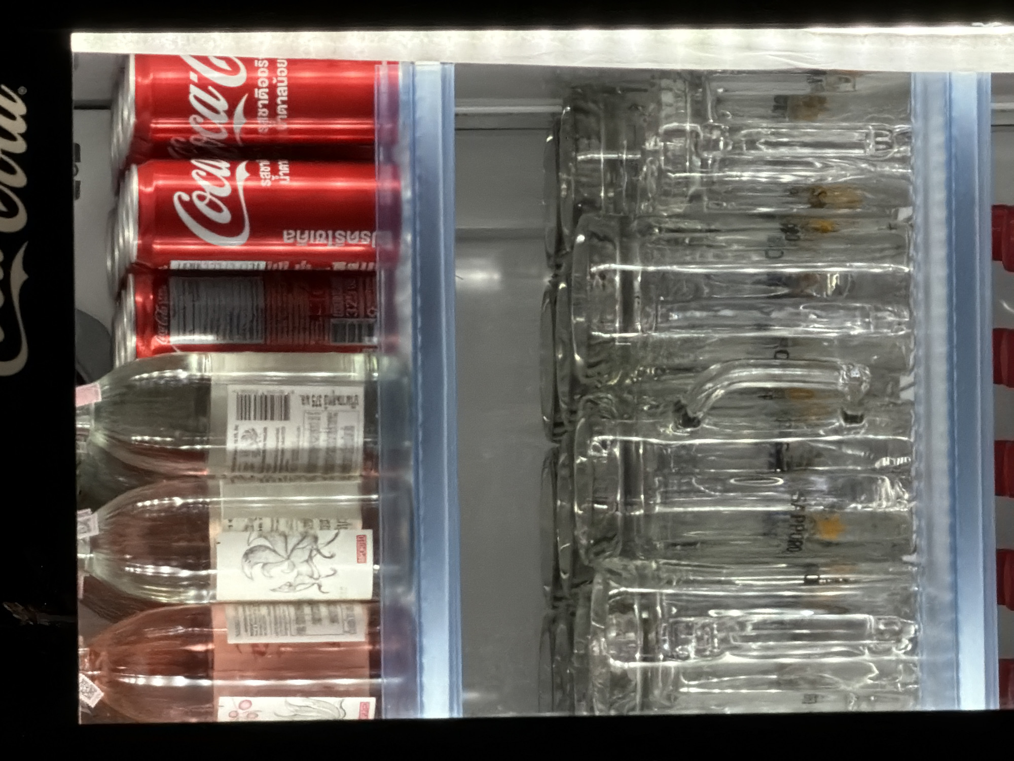 The image shows the inside of a commercial refrigerator. On the top shelf, there are several glass bottles containing a pink liquid, and to their right, there are red cans with white lettering that appears to be Coca-Cola branding. On the bottom shelf, there are rows of clear glass mugs. The refrigerator appears to be in a shop or restaurant setting. There are no people or animals visible in the image. The background is dark, and the lighting is focused on the refrigerator's contents. The image is taken from a slightly upward angle, looking into the refrigerator. The specific location within Bangkok, Thailand, cannot be determined from the visual information provided in the image.