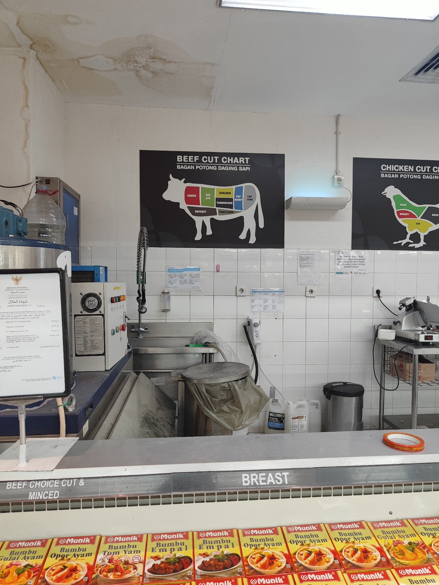 The image depicts a dimly lit interior of a food establishment, possibly a butcher shop or a supermarket meat section, in Nangkaan, Indonesia. The scene is dominated by stainless steel counters and tiled walls. On the far left, a certificate with Indonesian text is displayed. To its right, a large water dispenser with a red ribbon sits next to a piece of machinery, likely a refrigerator or a display unit, with several buttons and a fan. A faucet with a coiled hose is mounted above a stainless steel sink.

Dominating the background are two large black posters. The left poster features a "BEEF CUT CHART" with an illustration of a cow, detailing various cuts of beef in both English and Indonesian. The right poster displays a "CHICKEN CUT CHART" with a similar illustration and labeling in both languages. Above the posters, the ceiling shows signs of water damage, with discoloration and staining.

In the foreground, a refrigerated display case is filled with rows of packaged food items, predominantly labeled "Mamik" and featuring various Indonesian spice pastes ("Bumbu"). The words "BEEF CHOICE CUT & MINCED" are visible above this display, and "BREAST" is printed above a section of the display. To the right of the display case, a silver meat slicer is positioned on a metal shelving unit, which also holds some boxes and plastic containers. The overall environment suggests a commercial food preparation or retail area.