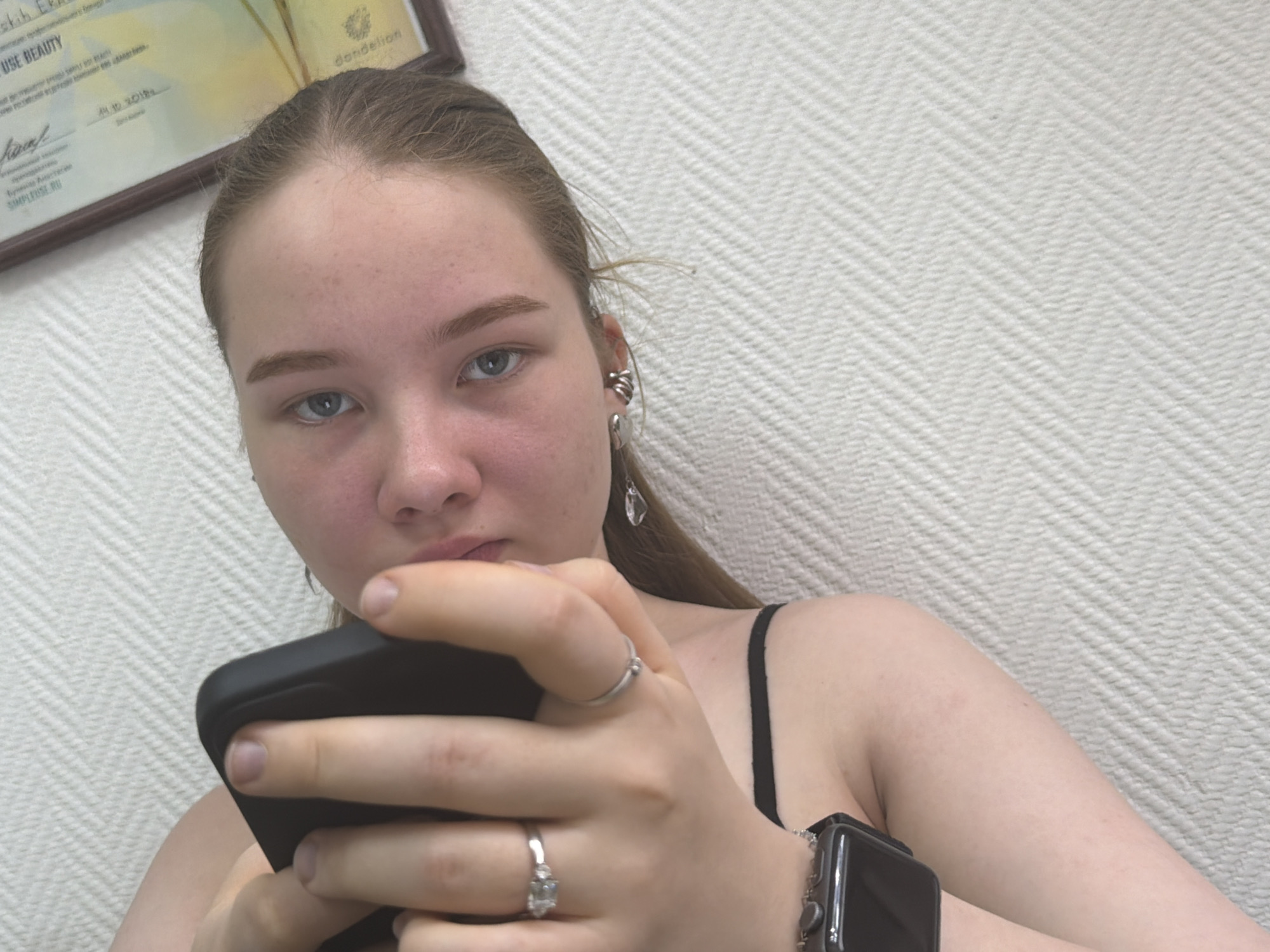 A young female subject is depicted in a head-and-shoulders, slightly low-angle portrait, looking directly forward. She has light brown hair, light blue eyes, and fair skin with some redness on her cheeks and nose. Her right ear is adorned with a silver ear cuff comprising multiple rings, a silver heart-shaped stud, and a dangling clear crystal earring. Her left hand, wearing a plain silver band on the ring finger and a silver ring with a clear stone on the middle finger, holds a black smartphone. A dark smartwatch with a metallic or beaded strap is visible on her left wrist. She is wearing a black strap garment. The background consists of a light-colored wall with a distinct herringbone pattern. In the upper left corner, a framed certificate is partially visible, displaying text such as "USE BEAUTY," "dandelion," "24.10.2018," and "Simplfyuse.ru," confirming the setting as Chaikovsky, Russia.