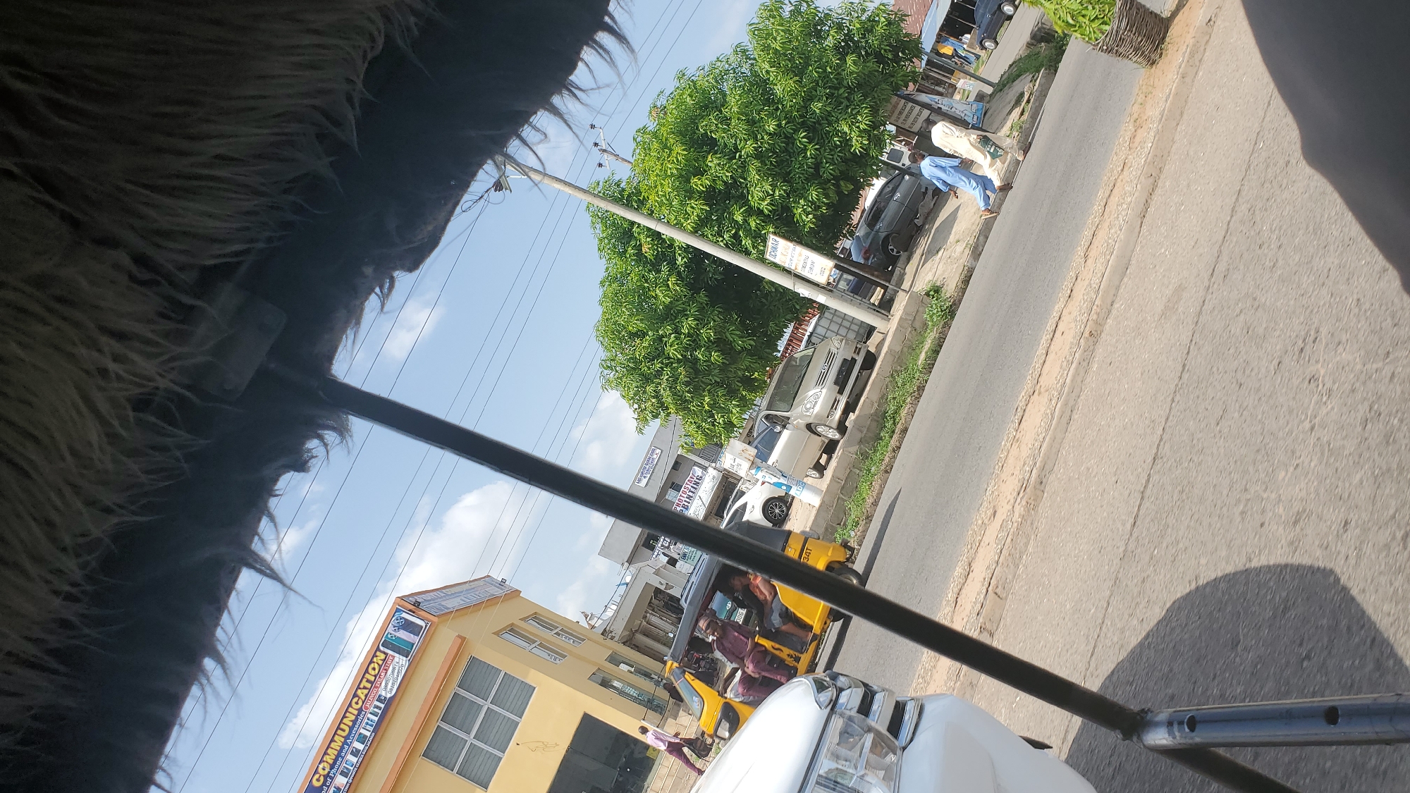 The image captures a street scene in Jalingo, Nigeria, as seen from a moving vehicle. In the foreground, a section of the vehicle's interior, including a fuzzy, dark-colored lining and a metal bar, partially obstructs the view. The street itself is paved, with a raised concrete curb running along its edge.
On the left side of the street, a two-story yellow building is visible. Its facade features several windows with prominent grilles. A sign on this building reads "COMMUNICATION" in large yellow letters, with smaller text beneath it mentioning "Head of Phone and Accessories" and listing a phone number. A person in a light pink shirt walks on the sidewalk in front of this building.
Further down the street, a yellow three-wheeled taxi, commonly known as a "Keke Napep," is transporting passengers. Two individuals are seated in the taxi, wearing traditional attire. A white car is parked on the right side of the street, partially visible in the foreground.
Across the street, a large, leafy green tree dominates the landscape, with a utility pole situated nearby. Several cars are parked along the sidewalk, and more buildings line the street in the background. Two men, dressed in light blue and cream traditional garments, are walking on the sidewalk. The sky is blue with scattered white clouds, indicating a daytime scene. The overall environment suggests a bustling urban setting with typical street traffic and commercial activity.