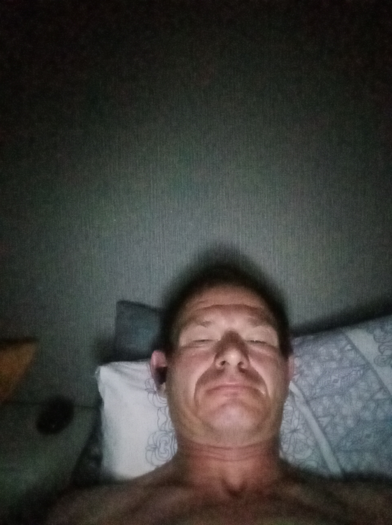 A man is lying down, likely in bed, as indicated by the pillow and blanket visible. He is looking up and towards the camera, with his eyes closed or partially closed, suggesting he might be tired or in the process of falling asleep. The lighting is dim, casting a shadow on the wall behind him. The wall has a textured pattern, and there is a hint of a darker object to his left, possibly furniture or another piece of bedding. The image is a close-up, focusing on the man's face and upper torso.