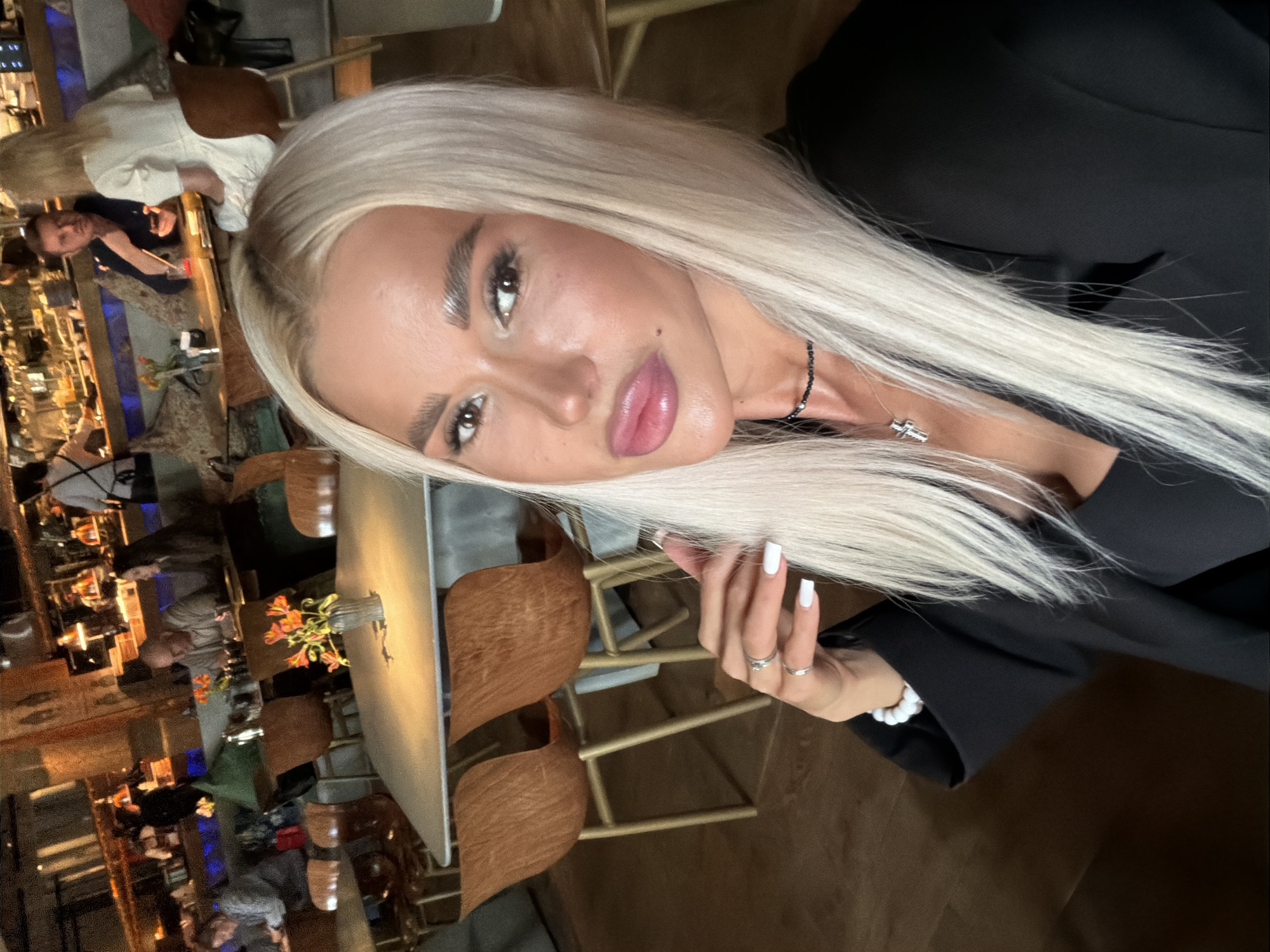 The image captures a close-up selfie of a woman with a blurred background suggesting an indoor dining or bar setting.
The main subject is a woman with very long, straight, light blonde hair that falls over her shoulders. She is looking directly at the camera with a slightly tilted head. Her facial features include well-defined, dark eyebrows, dark eye makeup, and full, glossy pink lips. A small mole is visible above her upper lip on the left side. She wears a black top or jacket, and a delicate necklace with a small, silver-colored cross pendant is visible around her neck. Another black beaded necklace or choker is also present. Her left hand, with white manicured fingernails and two silver rings (one on her index finger, one on her ring finger), is casually placed near her hair.
The background is out of focus but shows multiple people seated at tables, suggesting a social gathering or meal. The setting features warm, artificial lighting, indicating it might be evening or night. Unique wooden chairs with textured backs and light-colored tabletops are visible. A small vase containing orange flowers sits on one table in the mid-ground. Some blue lighting accents are discernible in the deeper background. No specific location cues or text are visible. The overall mood is relaxed and social.