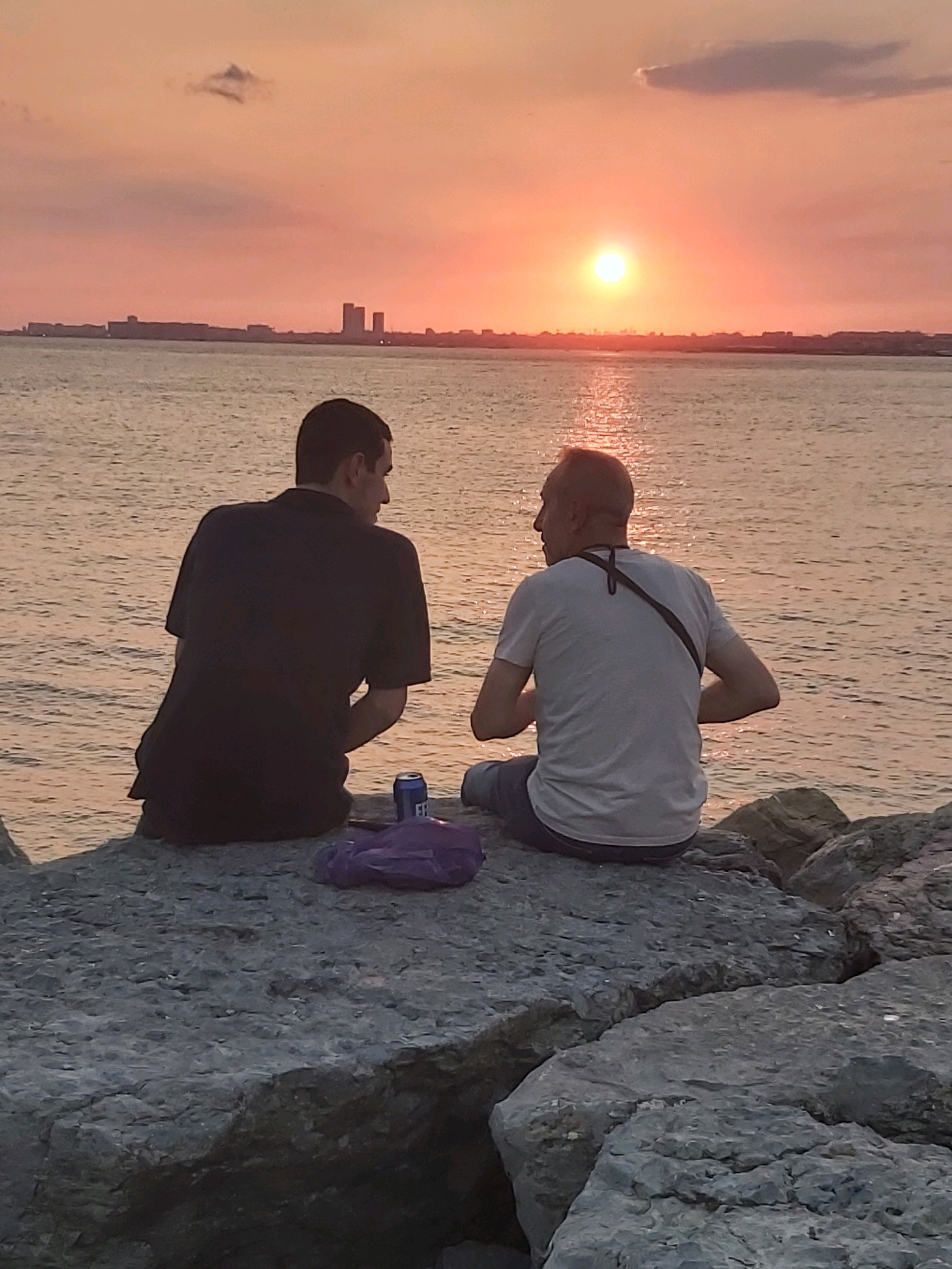 Two men are sitting on rocks by the sea, facing away from the camera, at sunset. The man on the left wears a dark t-shirt and the man on the right wears a light t-shirt. A purple bag and a can are on the rocks between them. In the distance, a city skyline with tall buildings is visible across the water, and the sun is setting over the horizon, casting an orange glow on the sky and water. The scene appears to be located in Kadıköy, Turkey, likely near the Haydarpaşa train station area, which features a similar waterfront and skyline.
