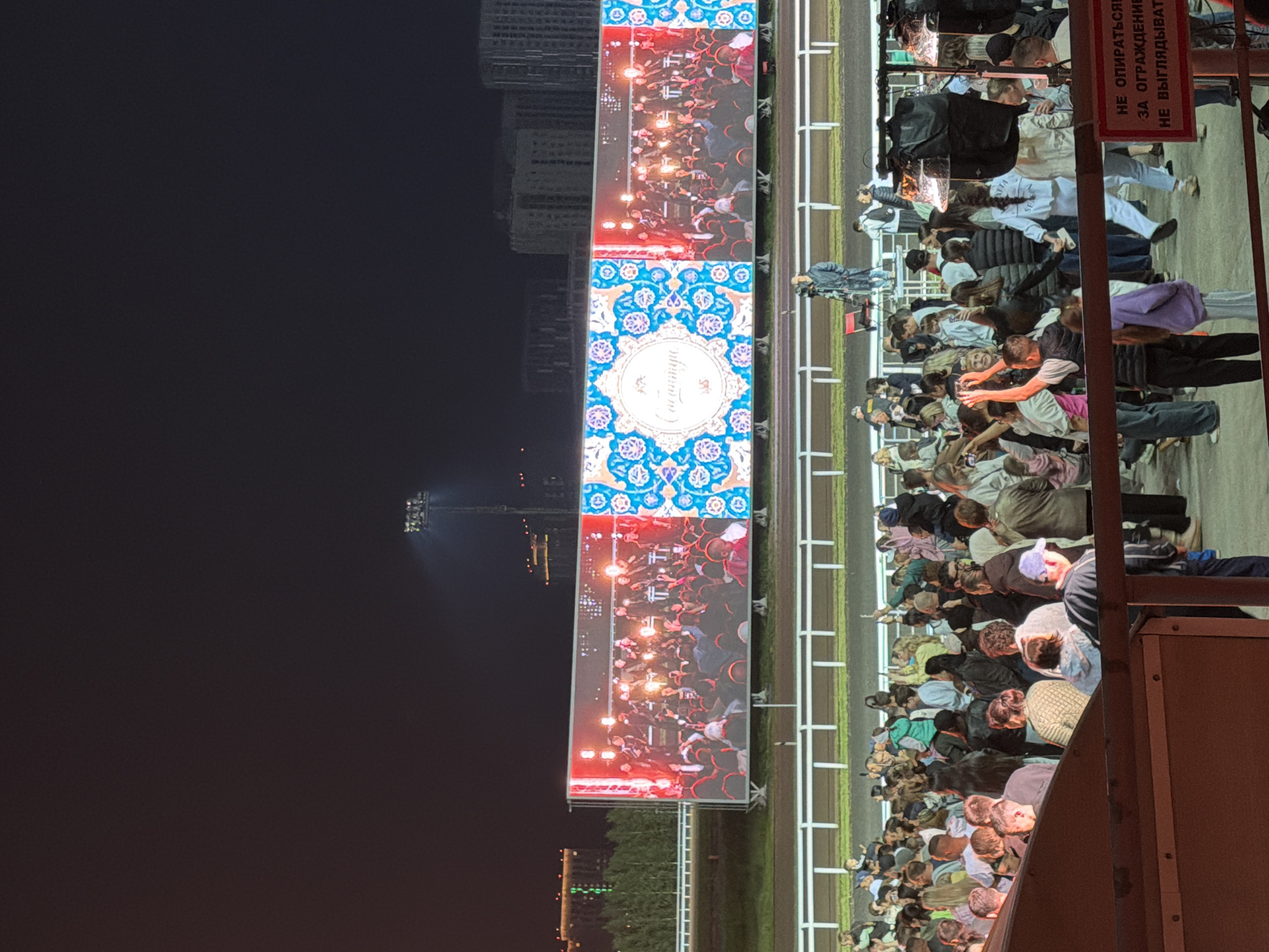 The image shows a large outdoor gathering of people at night. In the background, a large LED screen displays a concert or performance, with a crowd of people on stage and a decorative pattern on the left side of the screen. In front of the screen, a racetrack with white railings is visible, and more people are seated or standing behind it. The overall setting appears to be a stadium or event venue. The lighting is primarily from the screen and some stadium lights, creating a festive but dark atmosphere. The people in the foreground are a diverse group, some holding up phones, suggesting they are documenting the event.